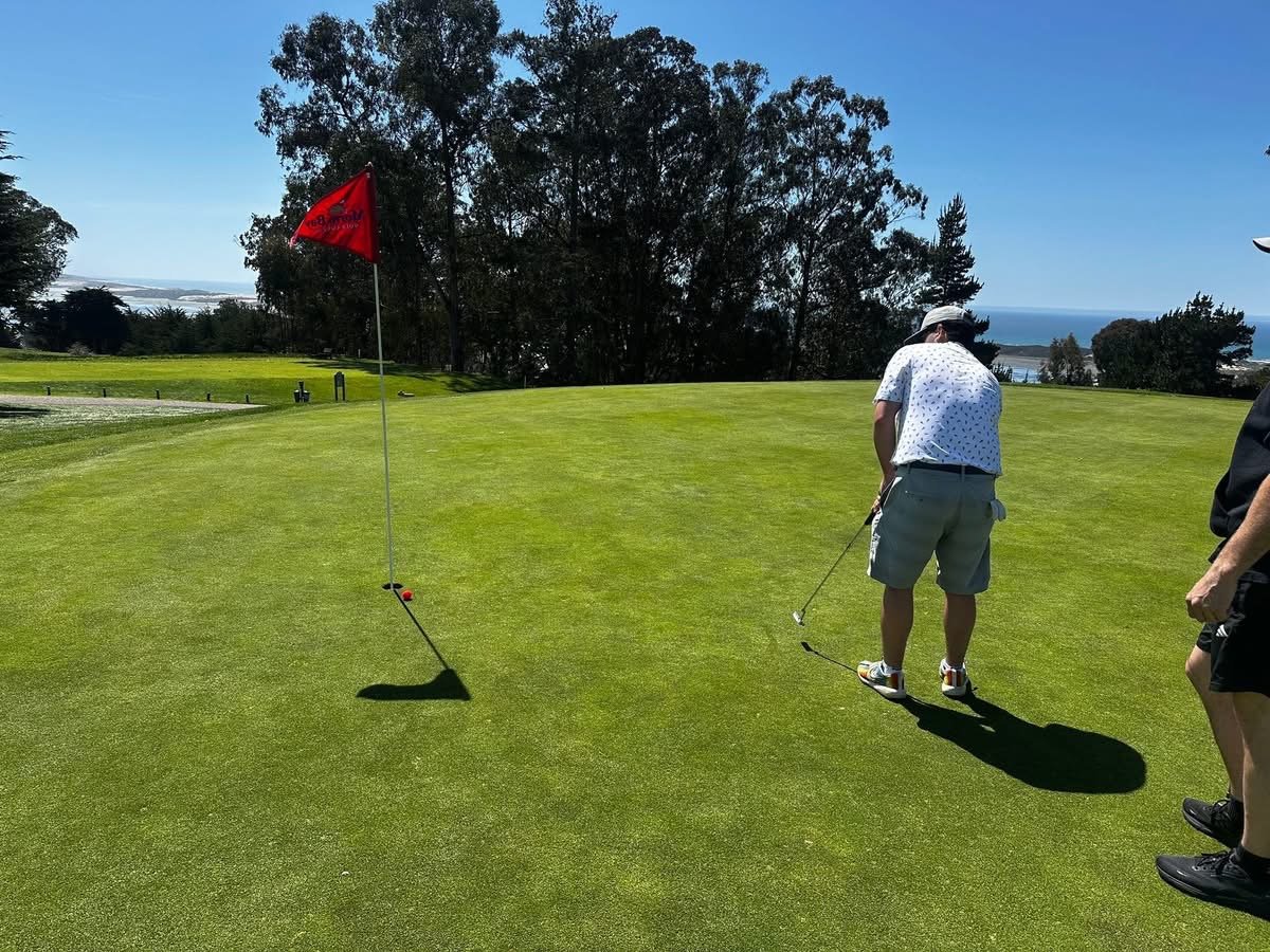Two men on a golf course near the green, with one preparing to putt and the other watching, under clear blue skies with trees and the ocean in the background.