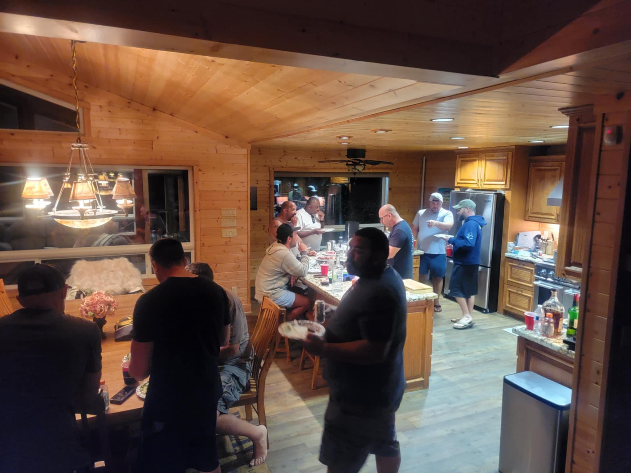 A group of people gathered in a wood-paneled kitchen and dining area, with some sitting at a table and others standing around the kitchen island, enjoying a social gathering or party.
