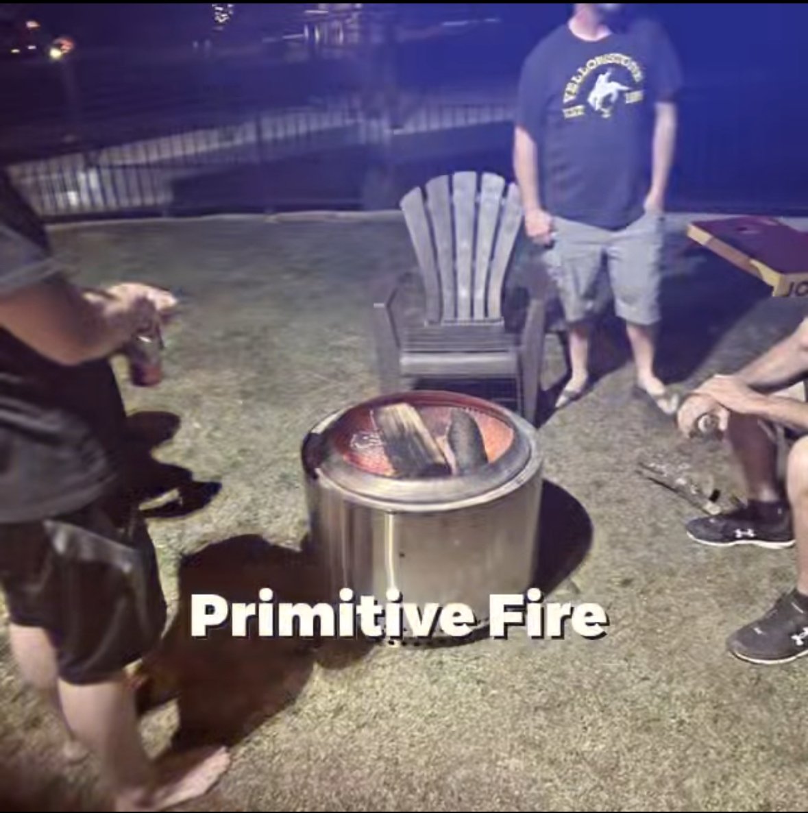 People gathered around a small fire pit on a grassy yard at night, with one person sitting on the ground, another standing, and a dog nearby, with the text overlay 'Primitive Fire'.