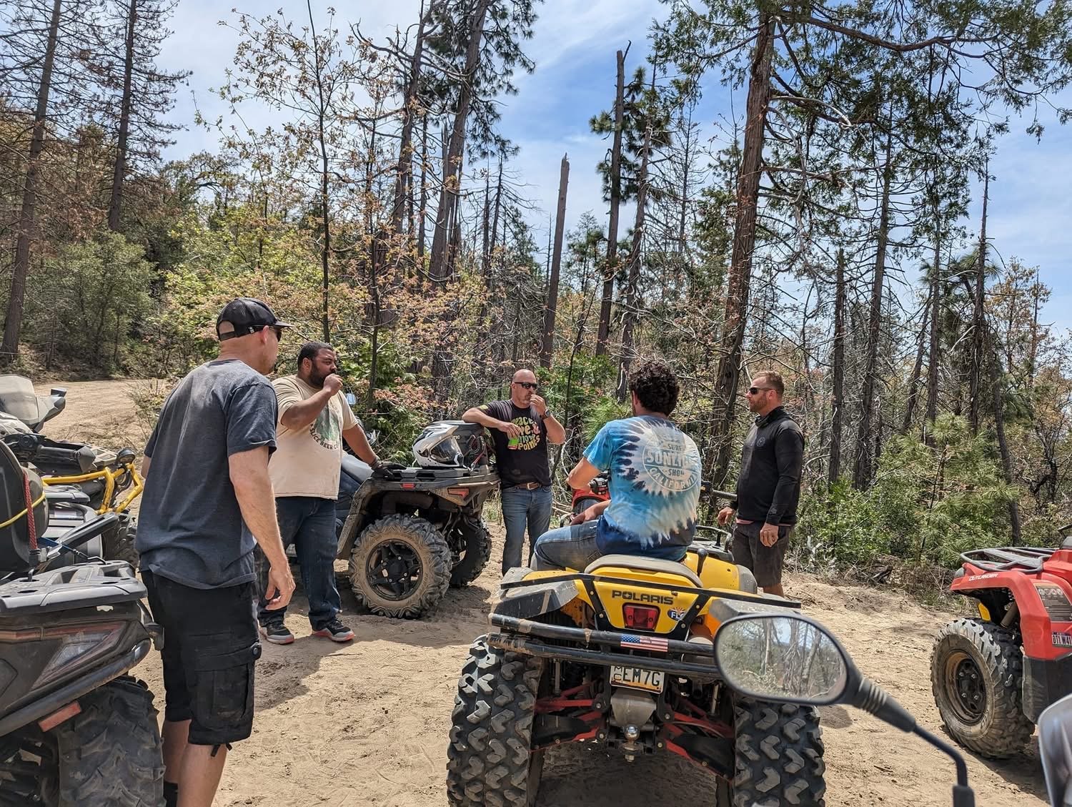 Group of six men gathered outdoors in a forested area with dirt ground, some sitting on all-terrain vehicles and others standing, engaging in conversation. The background shows tall trees and a clear sky.