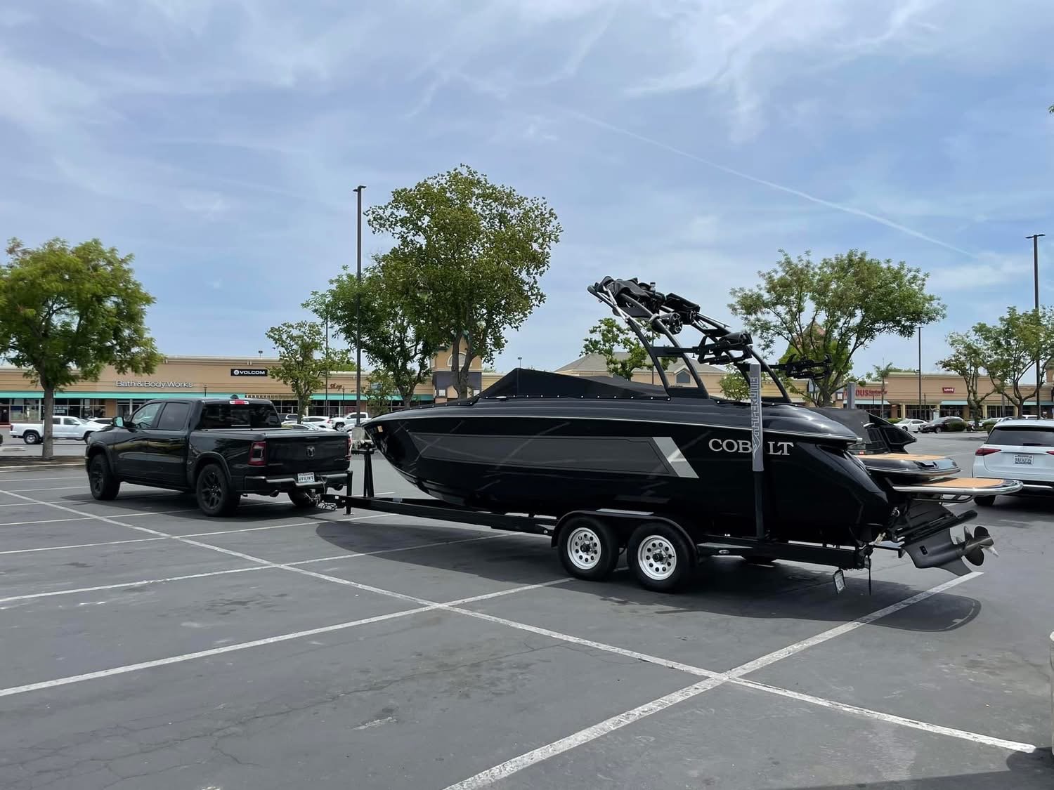 Black pickup truck towing a black boat on a trailer parked in a shopping mall parking lot, with stores and trees in the background.