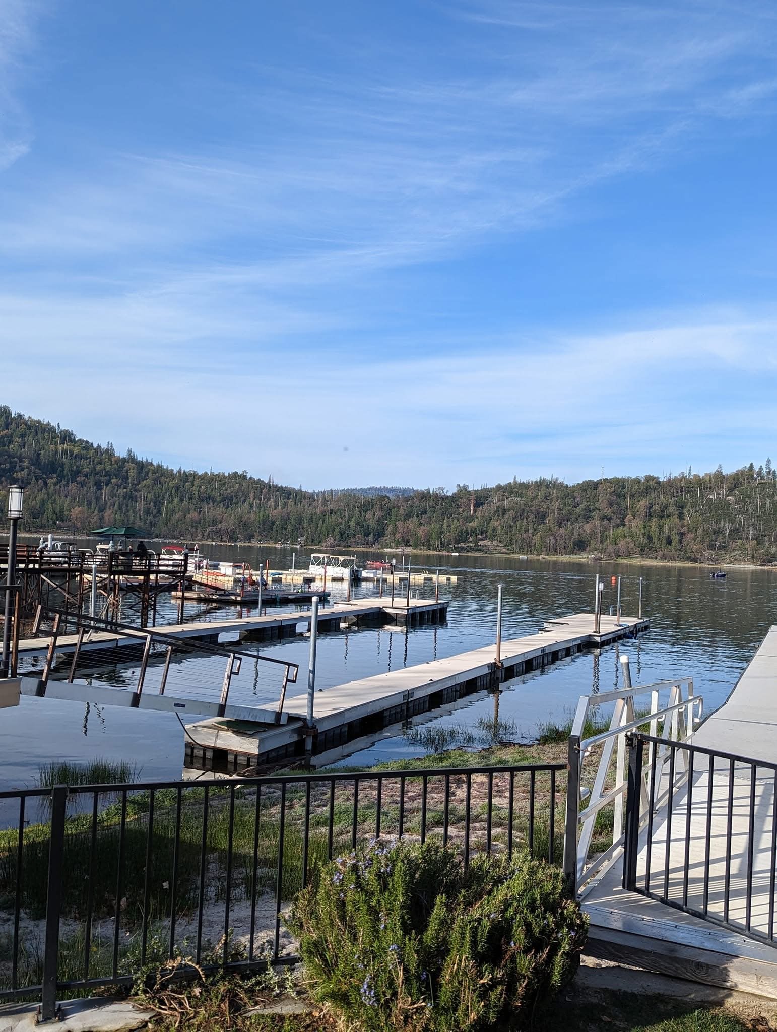 A scenic lakeside view with floating docks, a clear blue sky, hills covered with trees, and a walkway with a railing in the foreground.