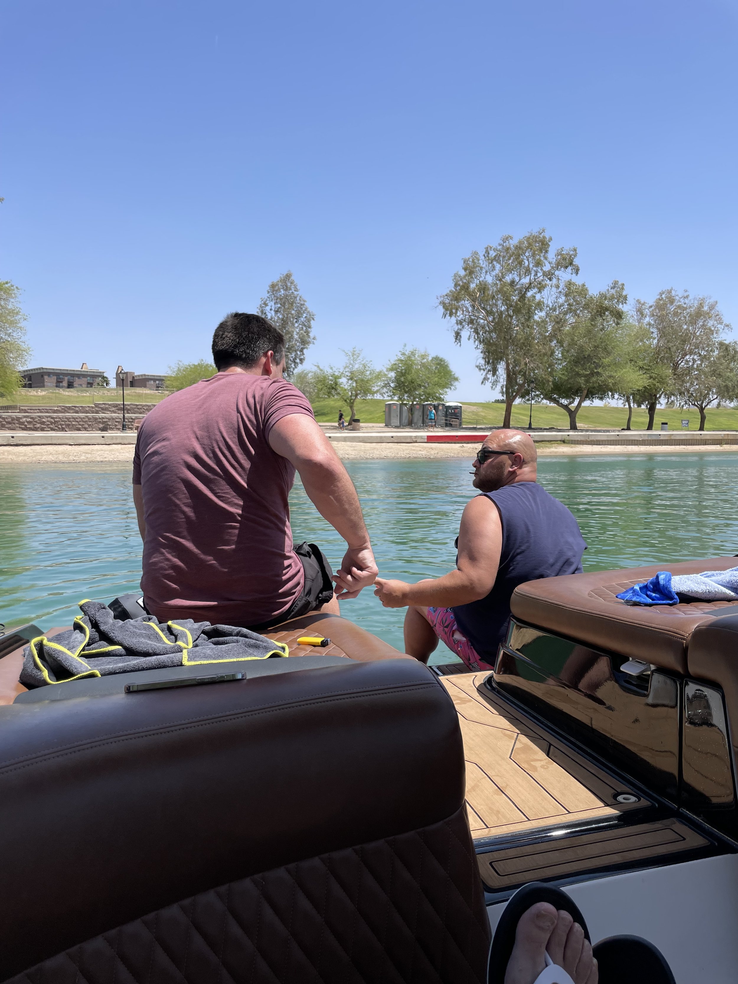 Two men sitting on the edge of a boat by a body of water, one wearing a maroon shirt and the other a navy tank top, holding hands, with trees and a clear blue sky in the background.