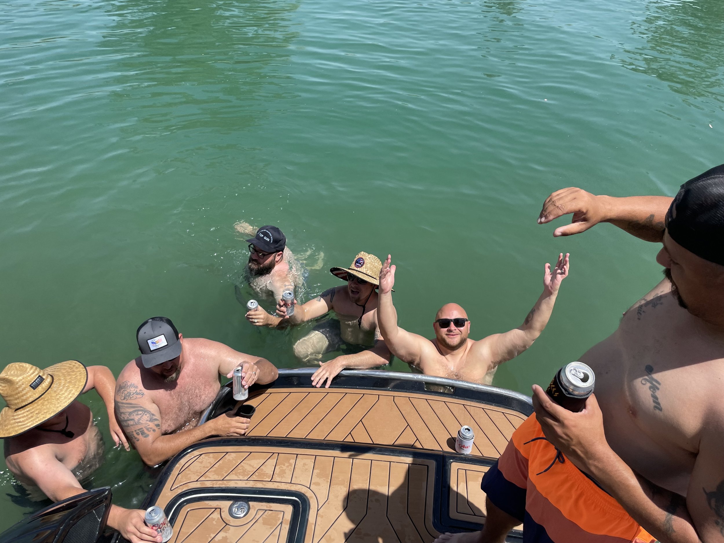 Group of men enjoying themselves at a boat party, some in the water, holding drinks, and making gestures.
