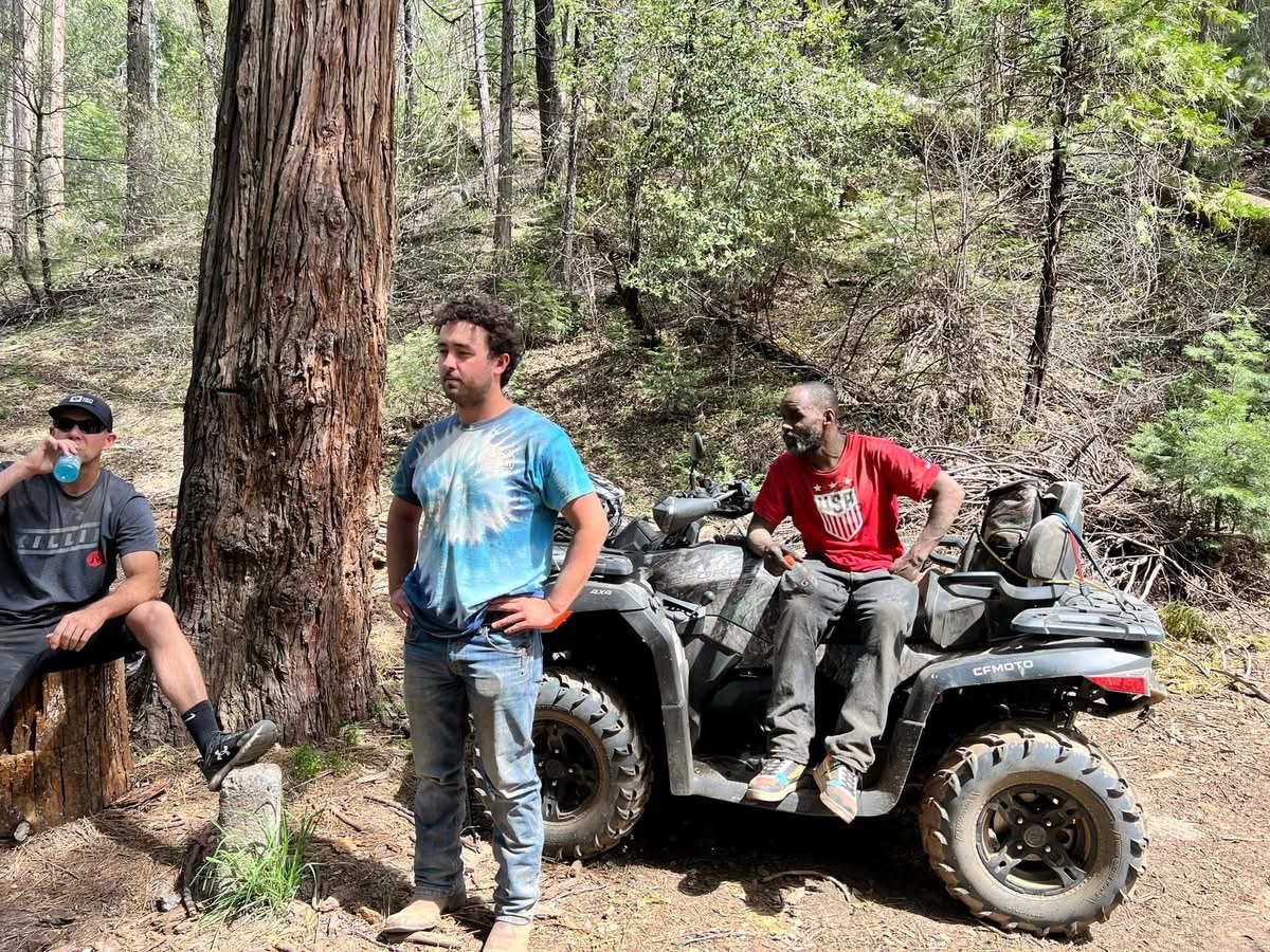 Three men in a forest, one sitting on a log drinking water, one standing with hands on hips, and one sitting on an all-terrain vehicle (ATV).
