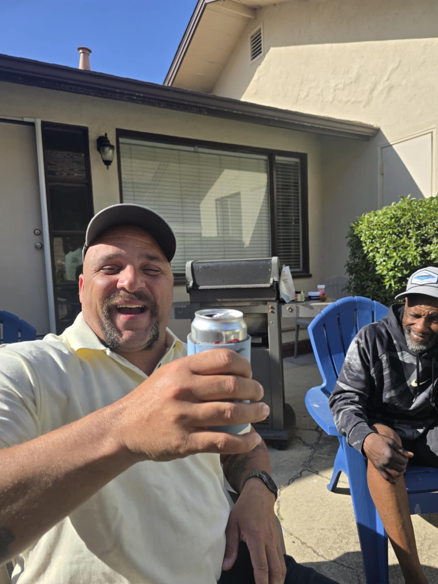 Two men sitting outside, smiling, with one holding a can of drink in the foreground.