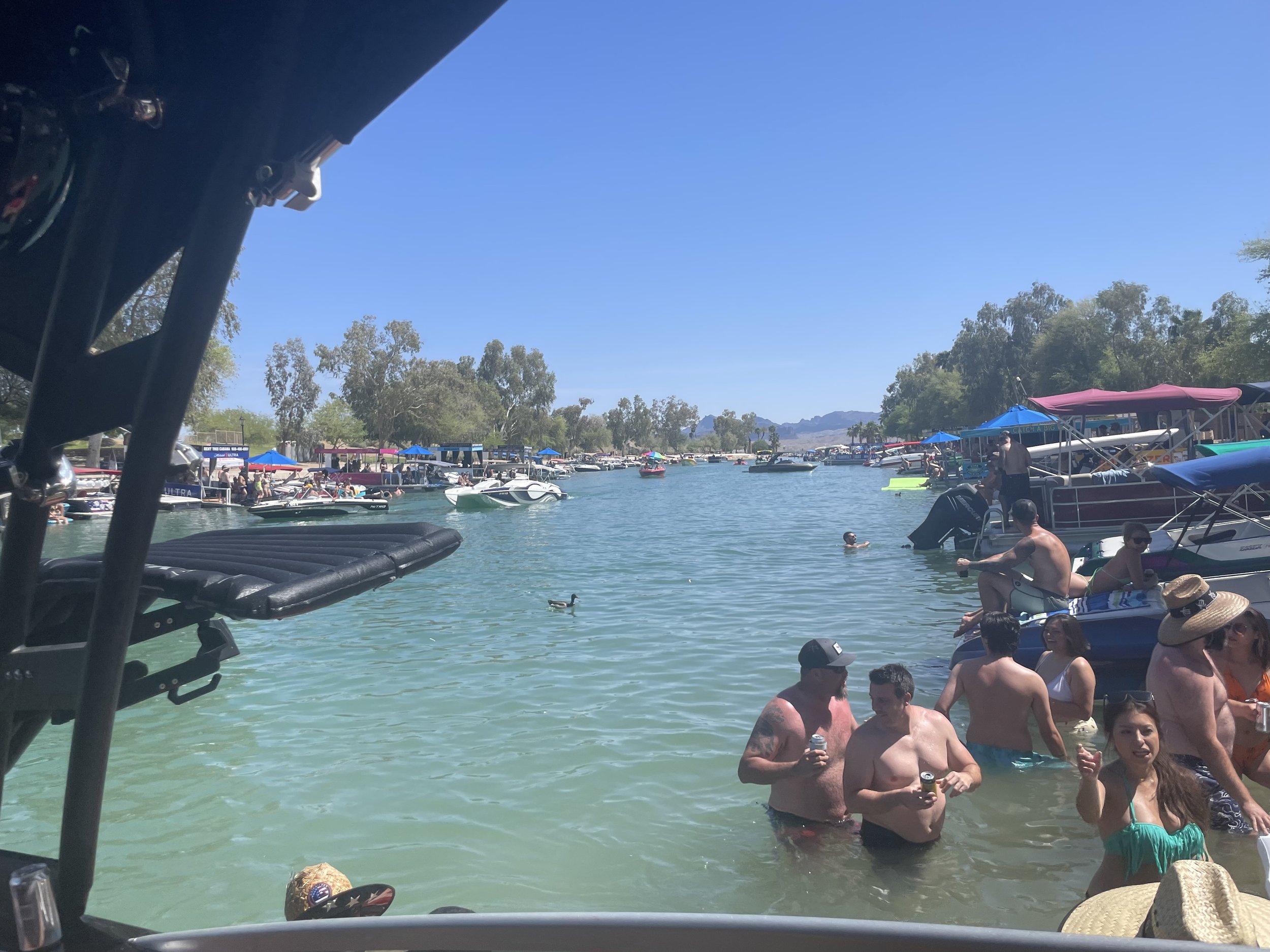 People swimming and relaxing in a crowded lake with boats and trees in the background on a sunny day.