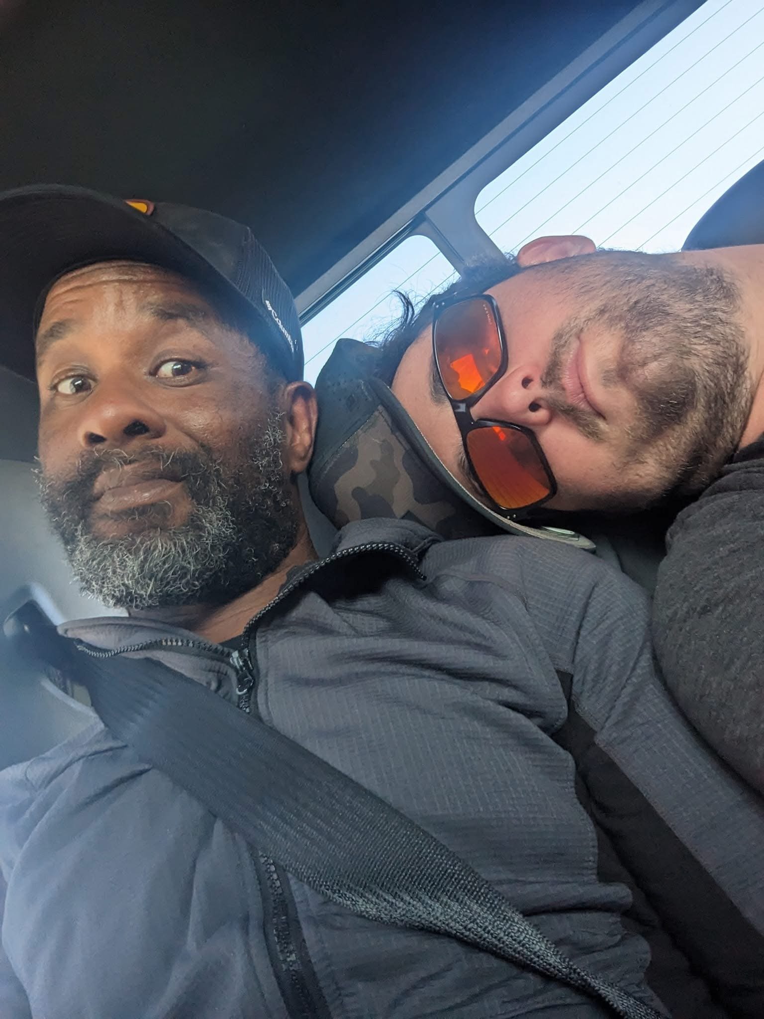 Two men sitting close on a vehicle, one with a gray beard and black cap, the other with sunglasses and dark hair, resting their heads together.