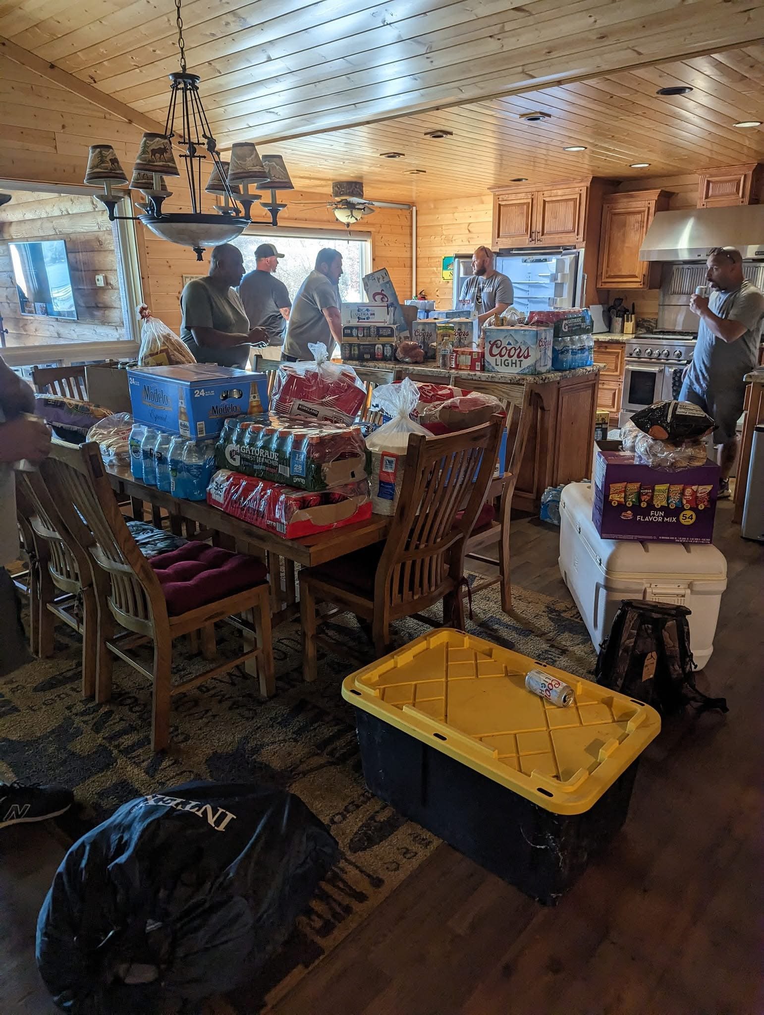 A group of people in a wooden kitchen, preparing for a gathering with tables full of food and drinks, and a large box of snacks nearby.