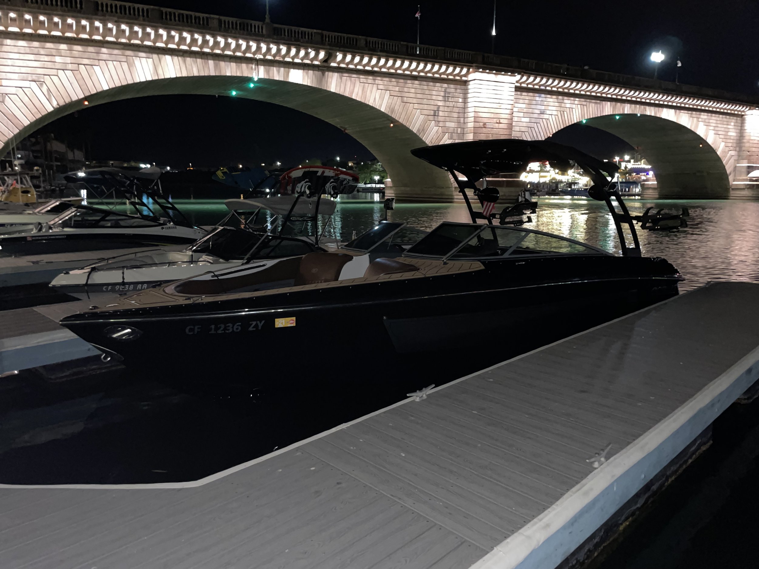 Nighttime scene at a marina with boats docked along a pier, a brick arched bridge overhead, and city lights reflected on the water.