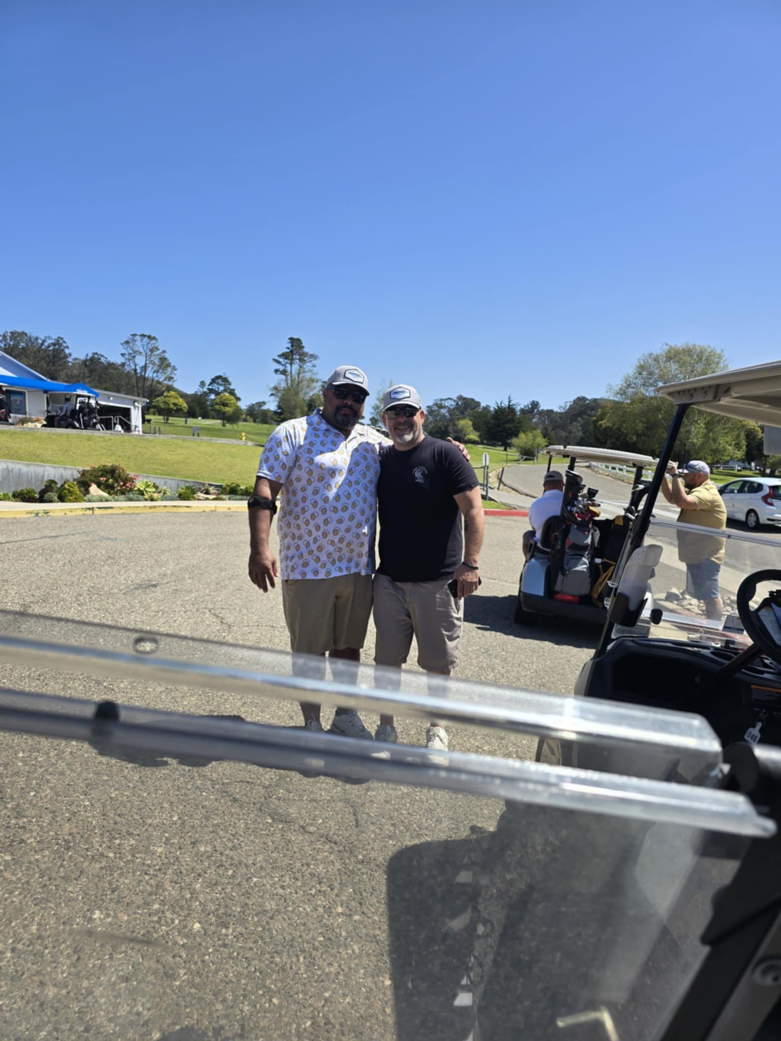 Two men standing side by side on a sunny day, near golf carts, with a grassy area and trees in the background.