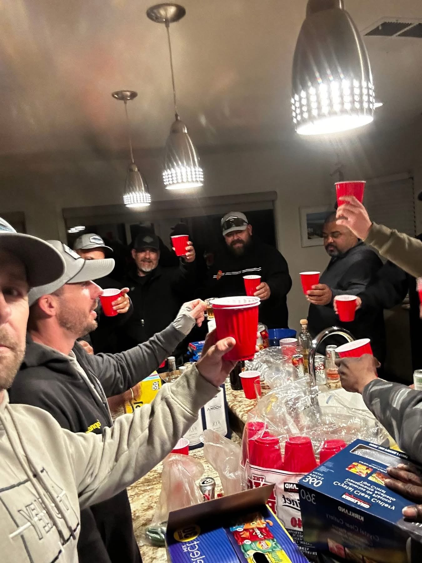 Group of people gathered around a kitchen island raising red party cups for a toast at a social gathering, with snacks and drinks on the counter.