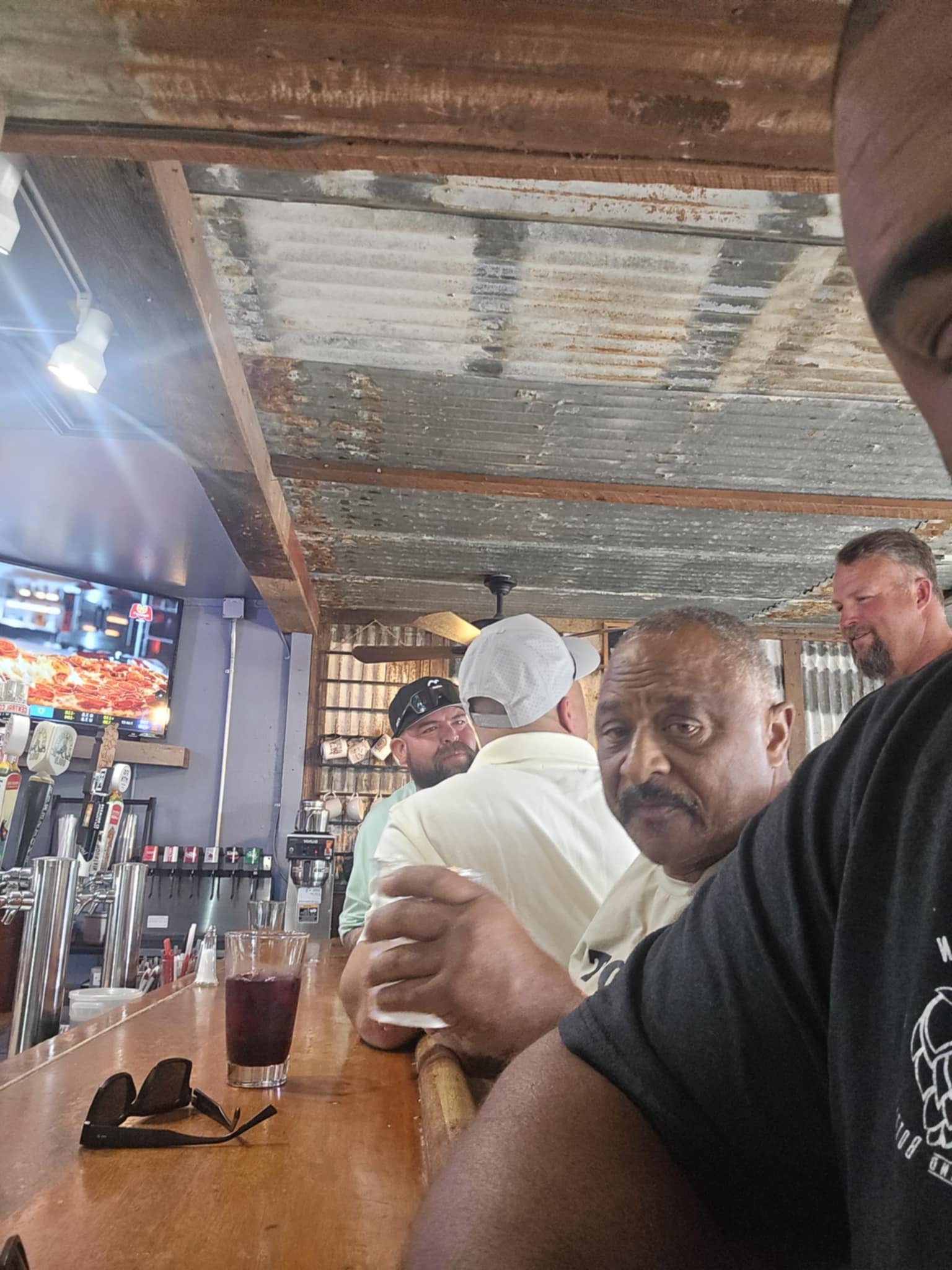 Man with a mustache holds a drink at a bar, with other men in casual clothing in the background, a television displaying pizza, and a rustic ceiling with metal and wood elements.
