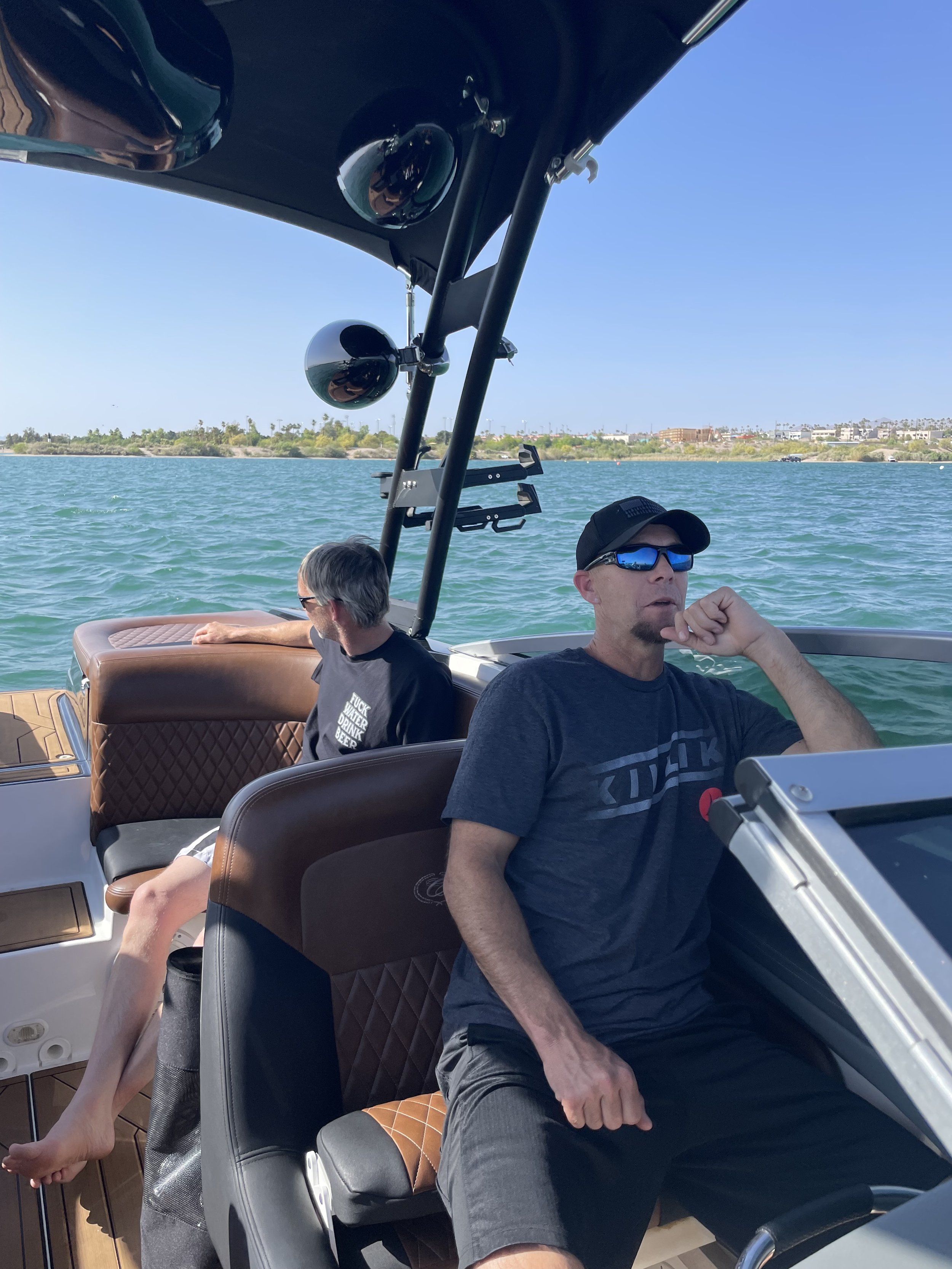 Two men on a boat on a sunny day, with water and distant shoreline in the background.