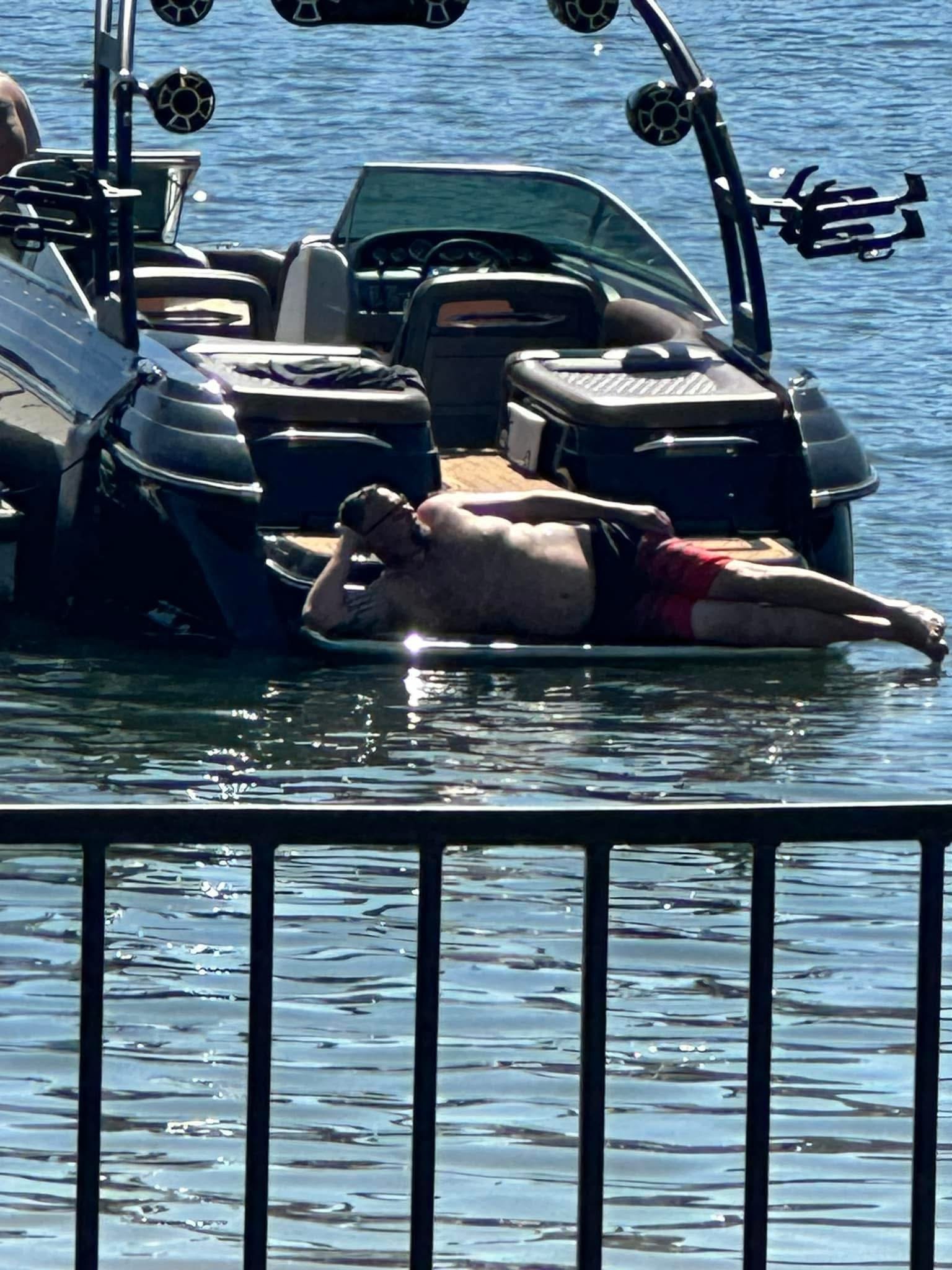A man lying on a boat docked at a waterbody, wearing red shorts and sunglasses, with his head propped up by one hand.