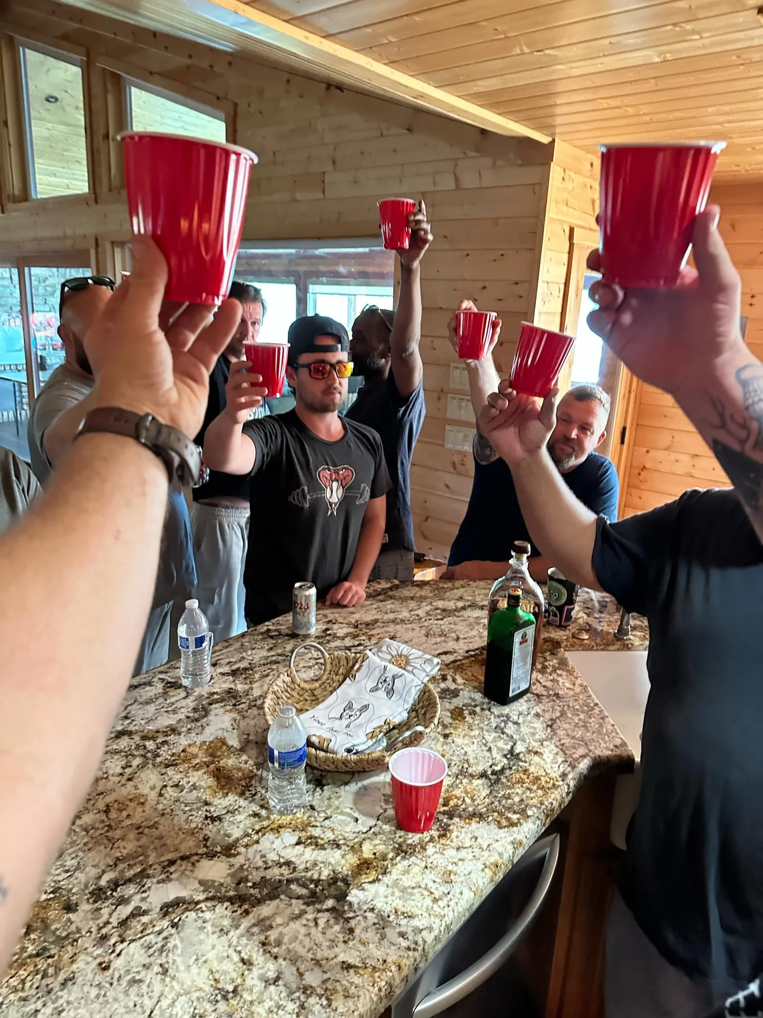 Group of people raising red plastic cups in a toast inside a wooden cabin kitchen.