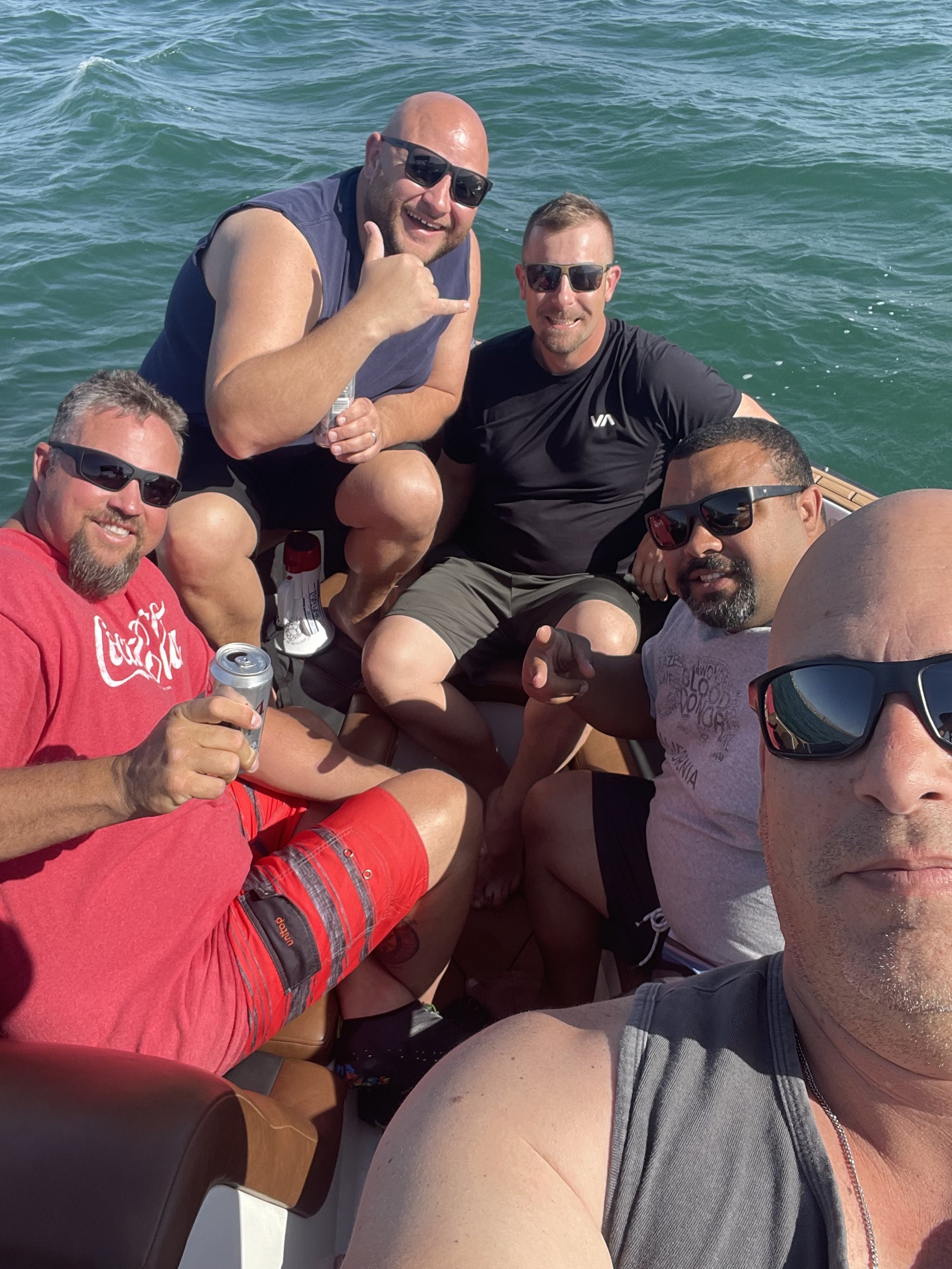 Group of six men on a boat enjoying the sun and water, some wearing sunglasses and casual summer clothes, with one man in the foreground taking a selfie.