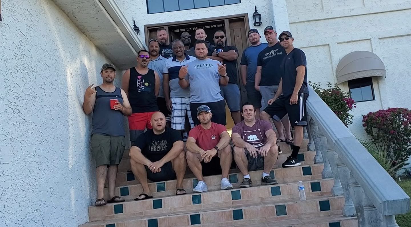 A group of 15 men gathered on the steps of a house porch, some sitting and some standing, smiling at the camera. The house has white stucco walls, a wooden door, and a glass window above it. There are flowers and bushes beside the staircase, and a wa