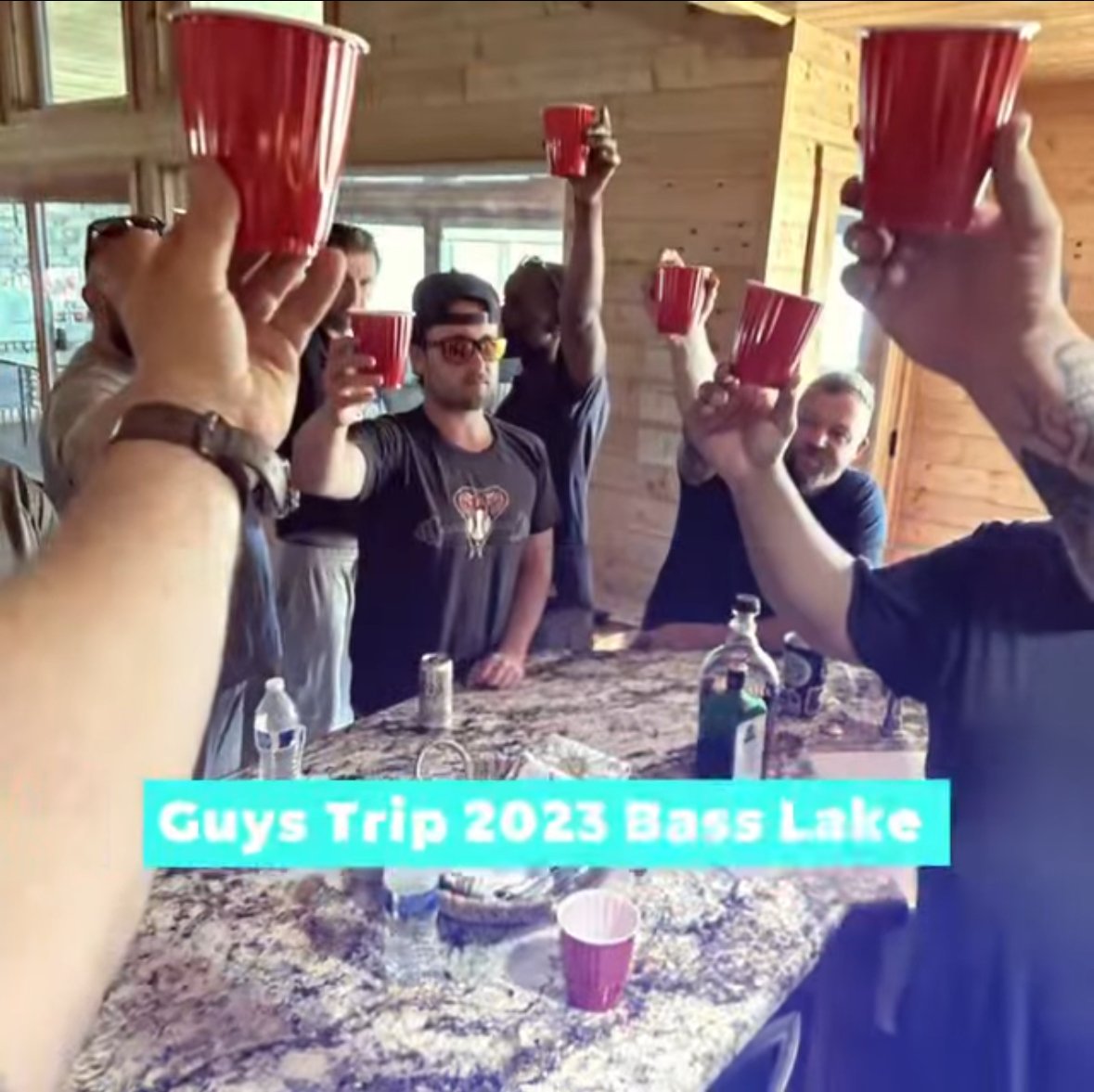 A group of people raising red cups in a toast during a trip to Bass Lake in 2023. They are inside a wooden cabin with a granite countertop in the foreground.