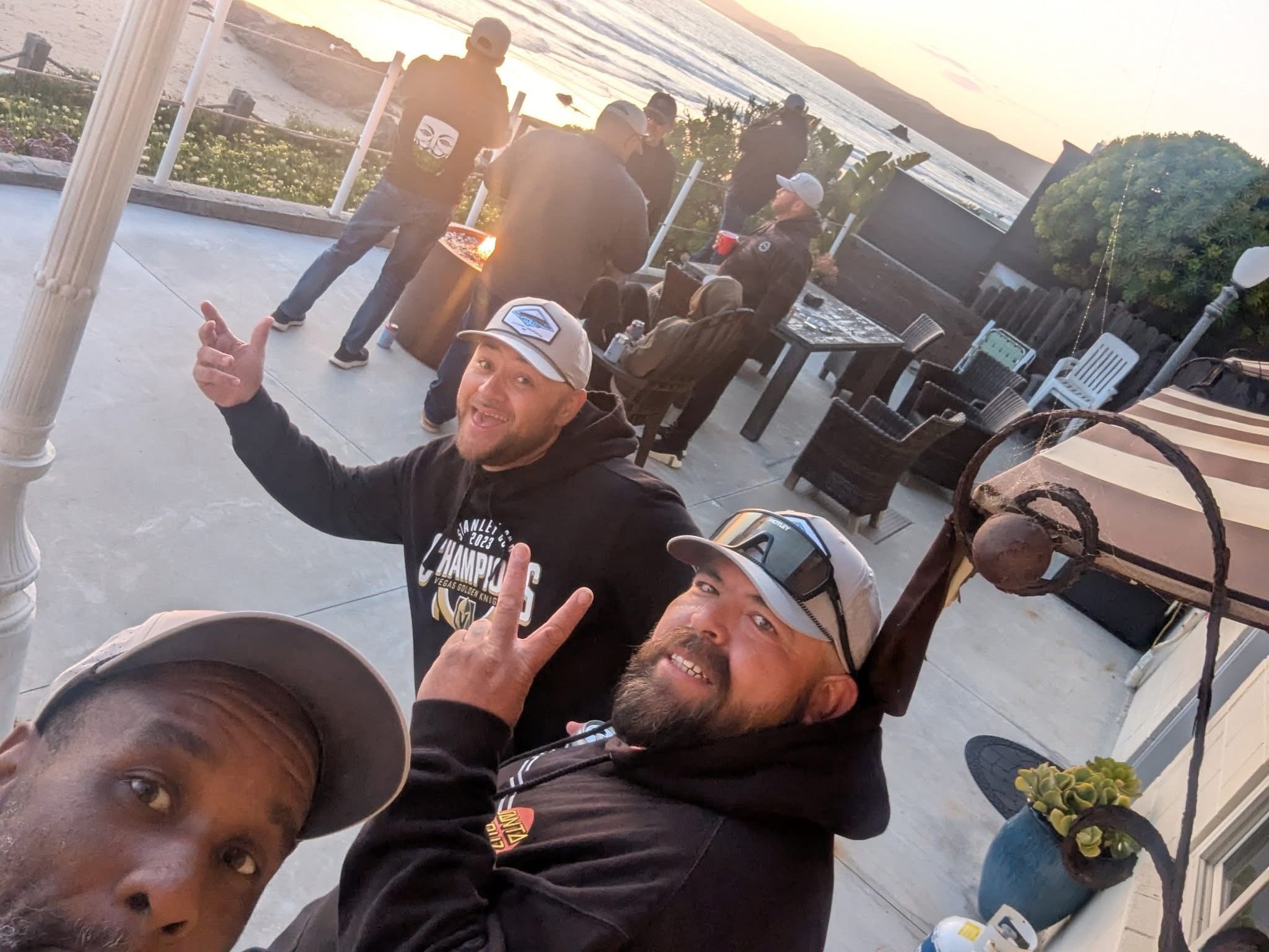 Group of friends enjoying outdoor barbecue at sunset by the ocean, some making peace signs and smiling.