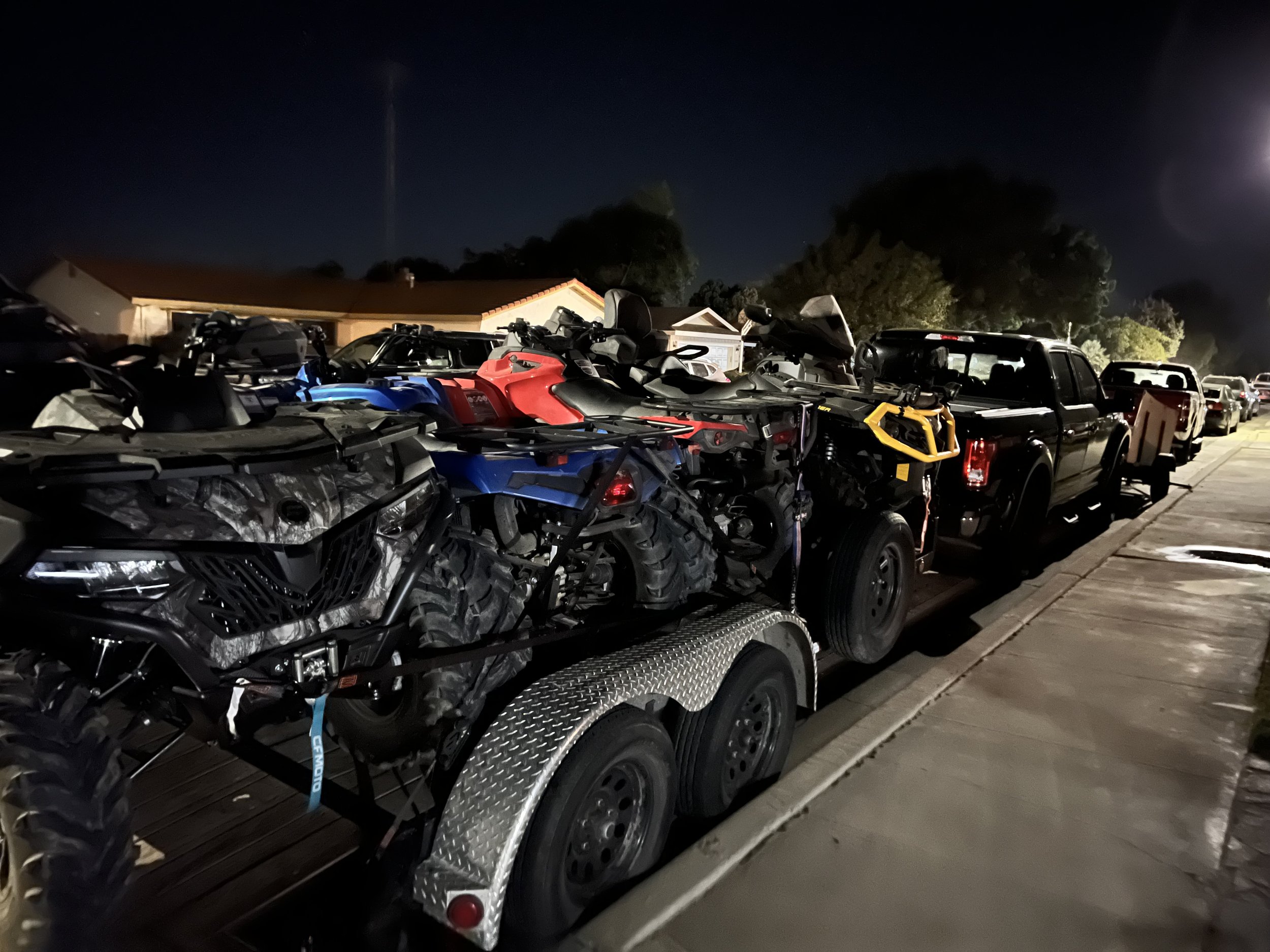 Nighttime scene of a trailer parked on a street loaded with multiple dirt bikes and ATVs, with cars parked along the curb and houses in the background.