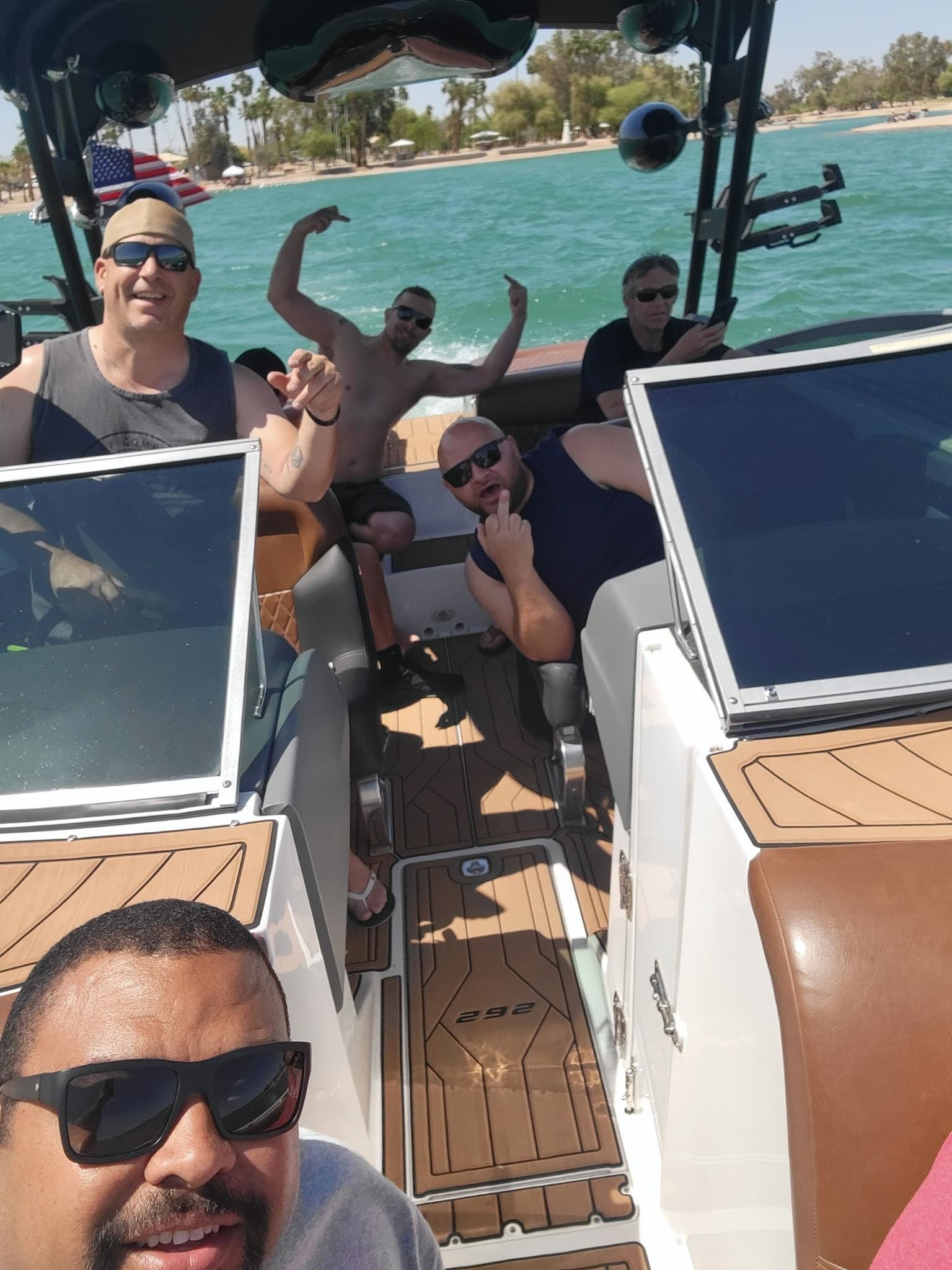 Six men are on a boat in a body of water with a beach and trees in the background. They are wearing casual summer clothes and sunglasses, with some smiling and making gestures.