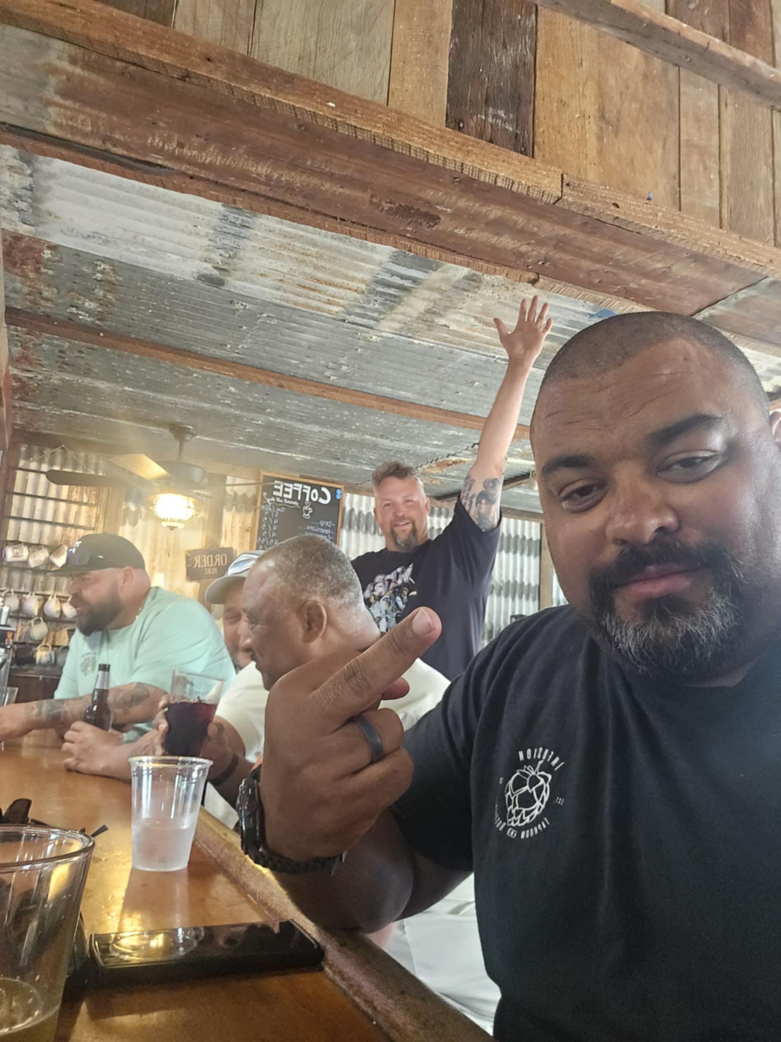 Group of men sitting at a bar, with one man in the foreground making a rude gesture. The man in the background is waving and smiling, wearing a black T-shirt. The bar has a wooden and rustic interior.