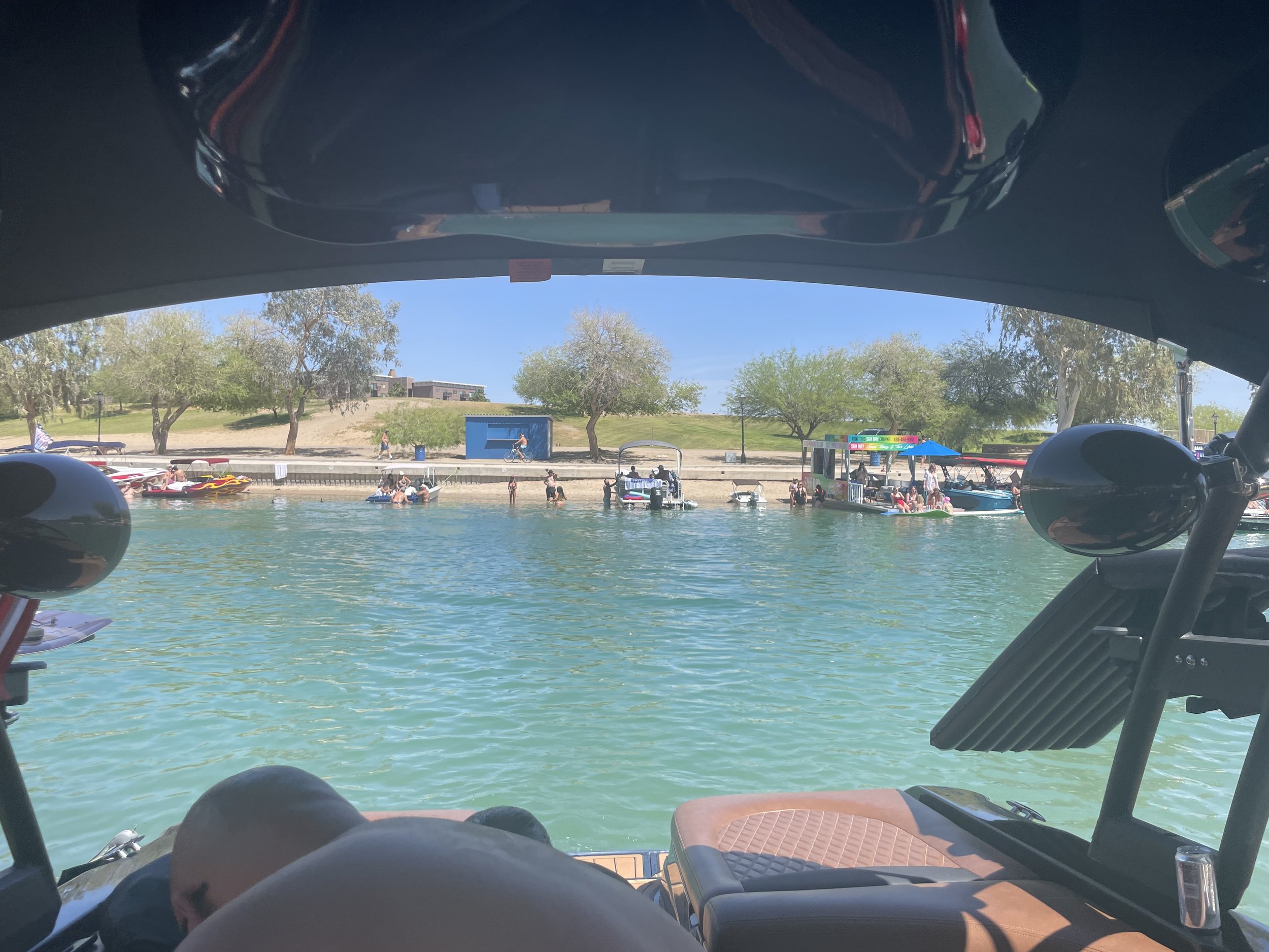 View from inside a boat looking out at the shore with people swimming and boats docked along the water's edge, trees, and a grassy hill in the background.