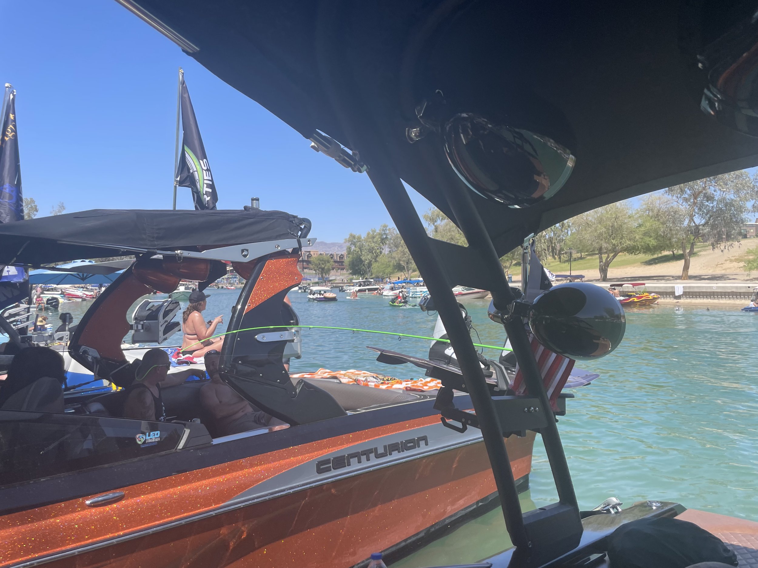 View of a boat with a glittery orange and black design on a calm waterway, seen from beneath a canopy with sunglasses hanging on a frame, with people on the boat and other boats and trees in the background under a clear blue sky.