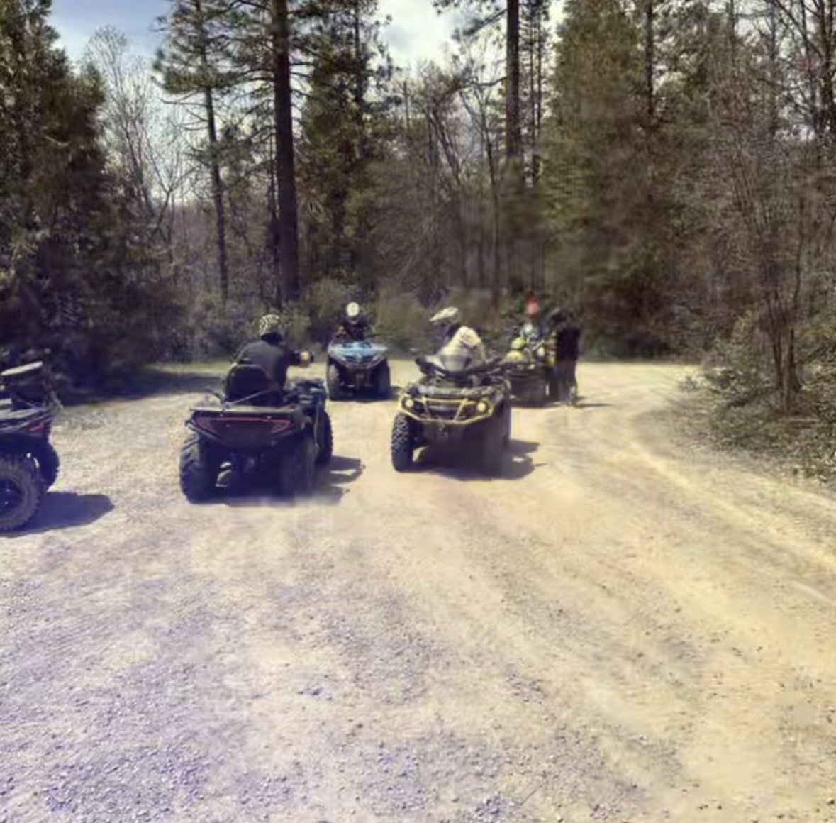 Group of people gathered around ATVs in a wooded outdoor area, some wearing helmets, on a dirt path.