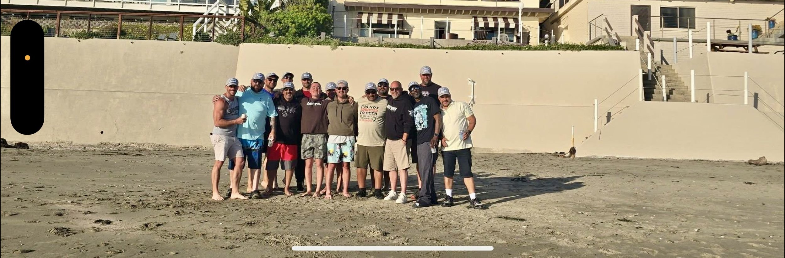 A group of around 15 men standing on a sandy beach in front of a concrete wall, with some houses and stairs visible in the background. Most of the men are wearing casual clothes, sunglasses, and baseball caps. Some are holding drinks, and one person