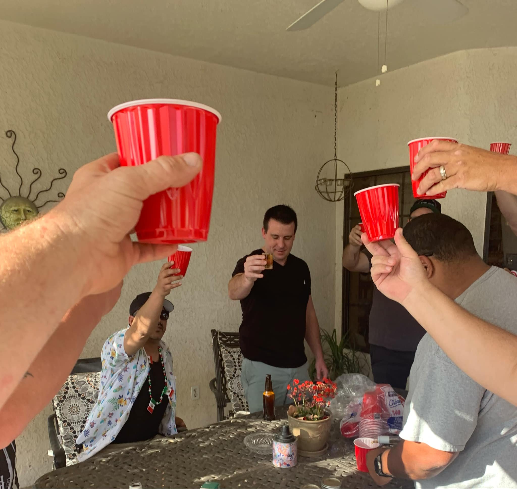 Group of people raising red striped plastic cups in a toast during a celebration at an outdoor table with food and drinks.