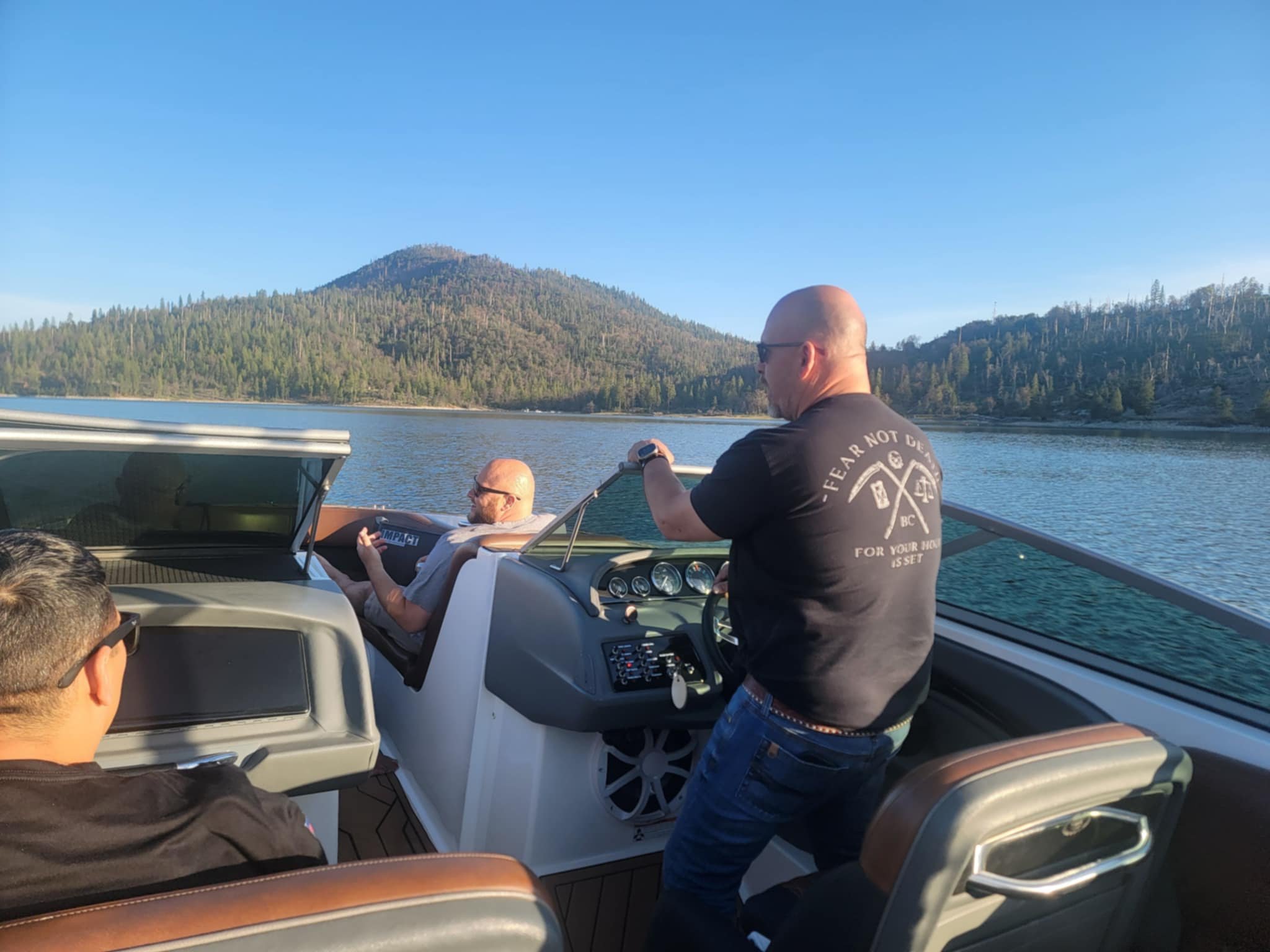 Three men on a boat on a lake with mountains in the background, one man is steering the boat, another man is sitting and using a phone, and the third man is sitting and wearing sunglasses.