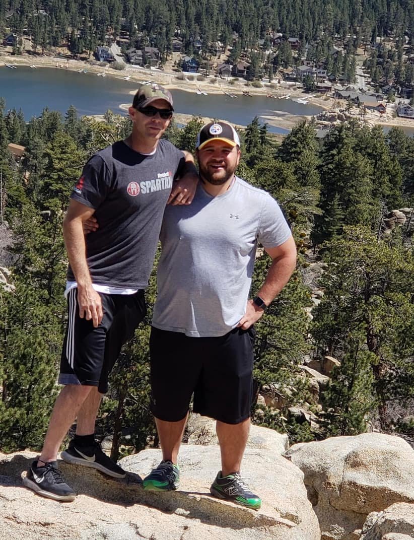 Two men standing on rocks outdoors, with trees, a lake, and houses on a hillside in the background.