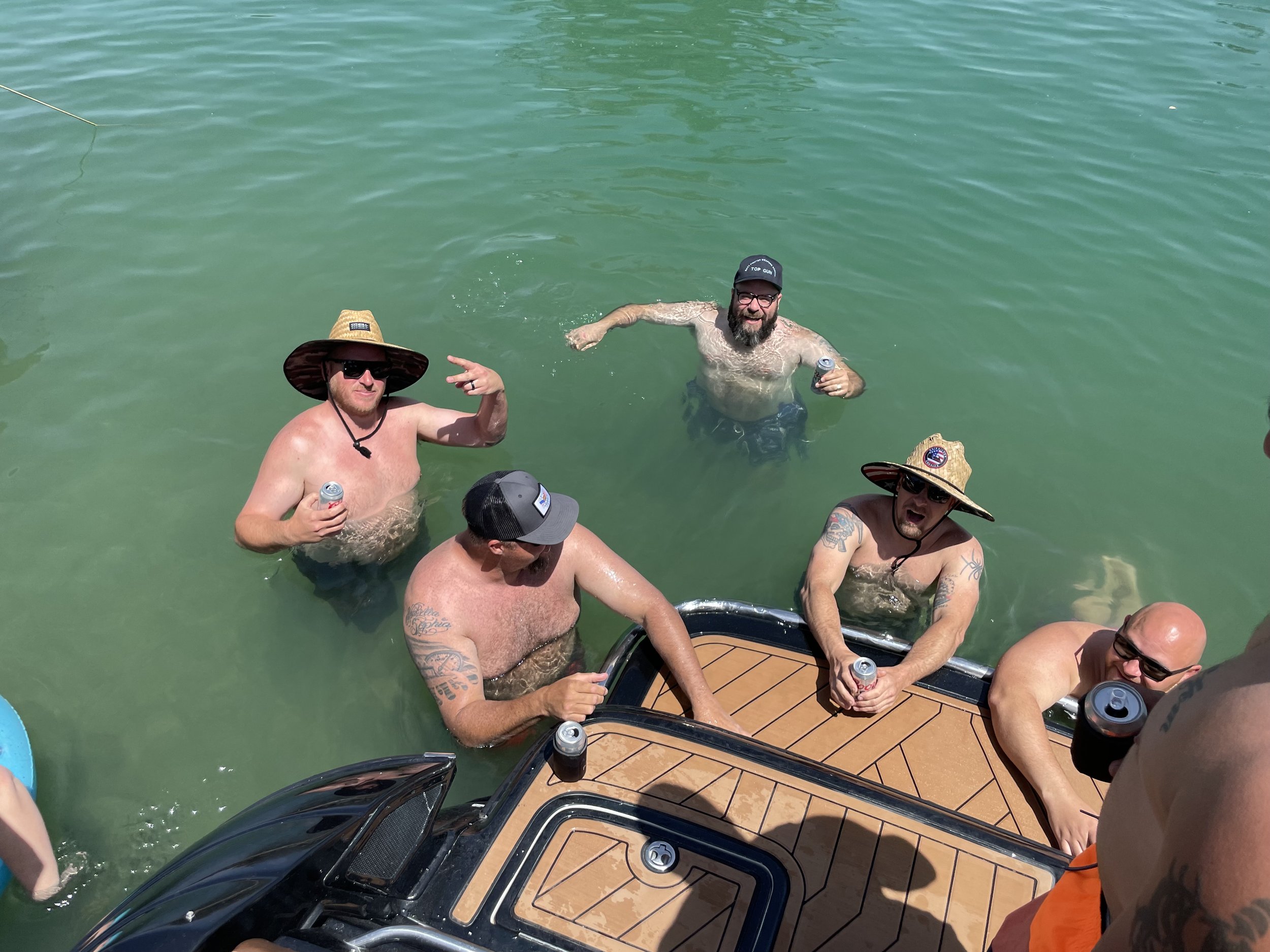 Group of five men in swimwear and hats enjoying a swim and drinks near a boat in green water