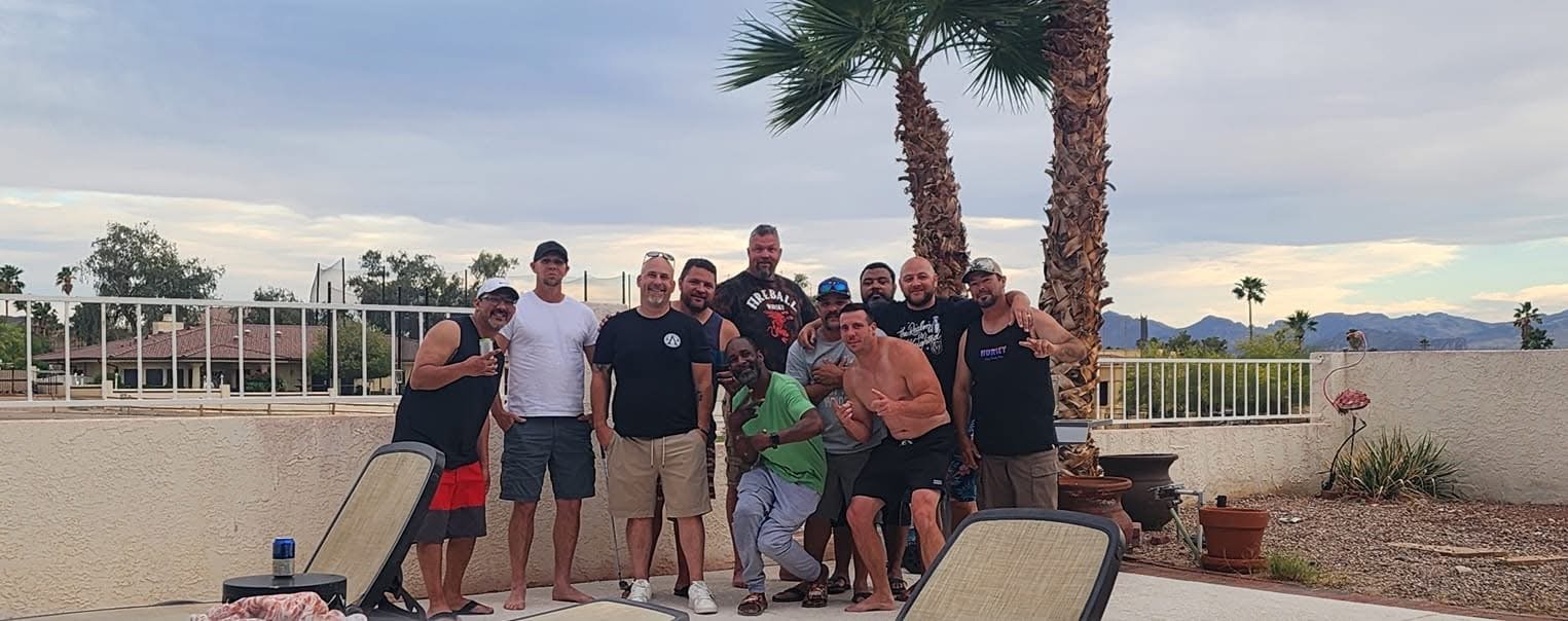 Group of men posing outdoors near a pool, with palm trees and mountains in the background.