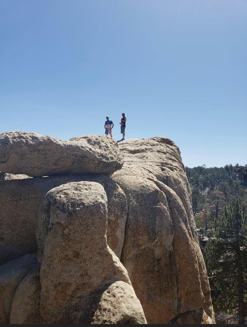 Two people standing on large rocks at a high vantage point with a clear blue sky and trees in the background.