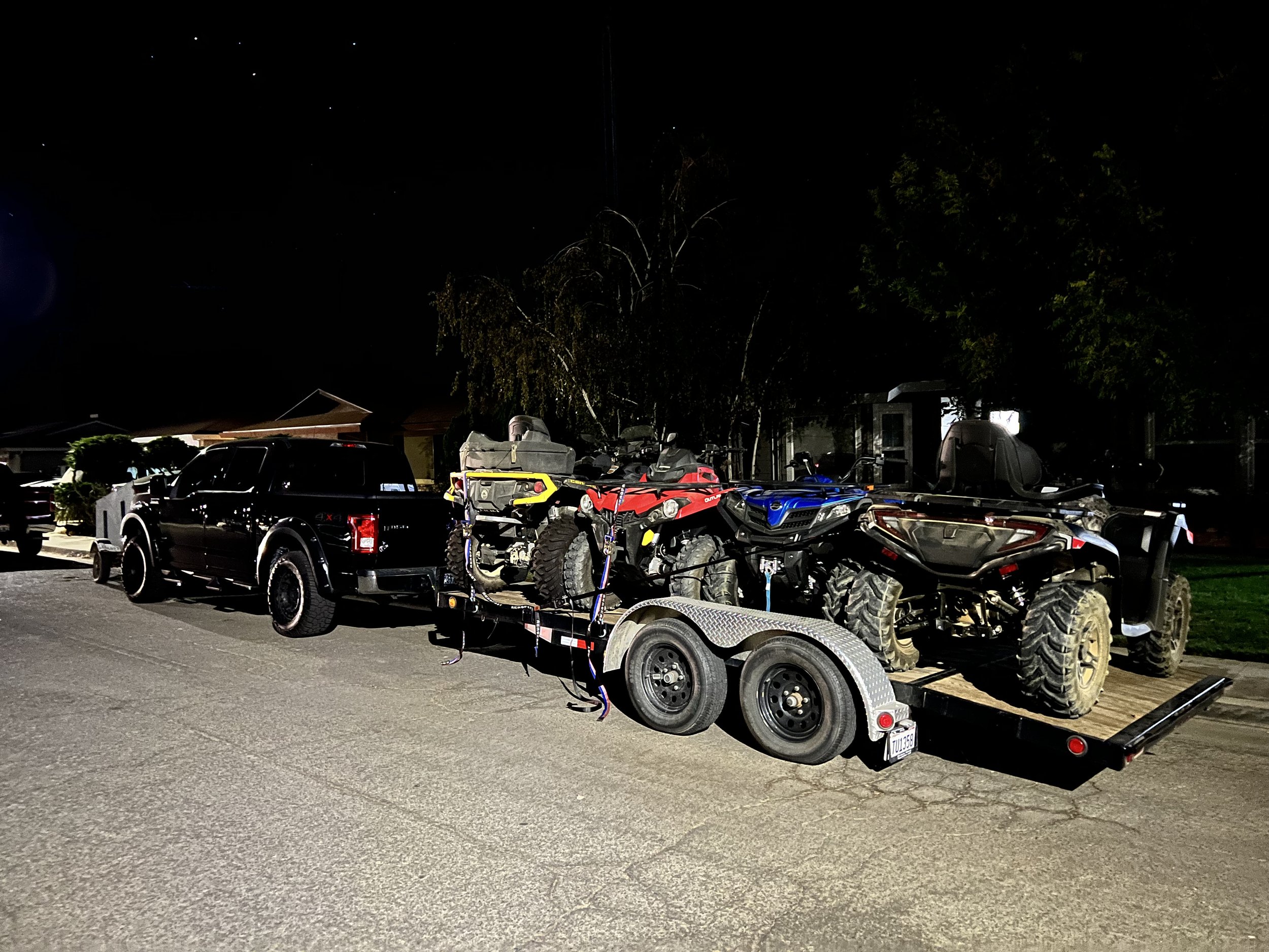 Black pickup truck with a trailer carrying four all-terrain vehicles (ATVs) parked on a residential street at night.