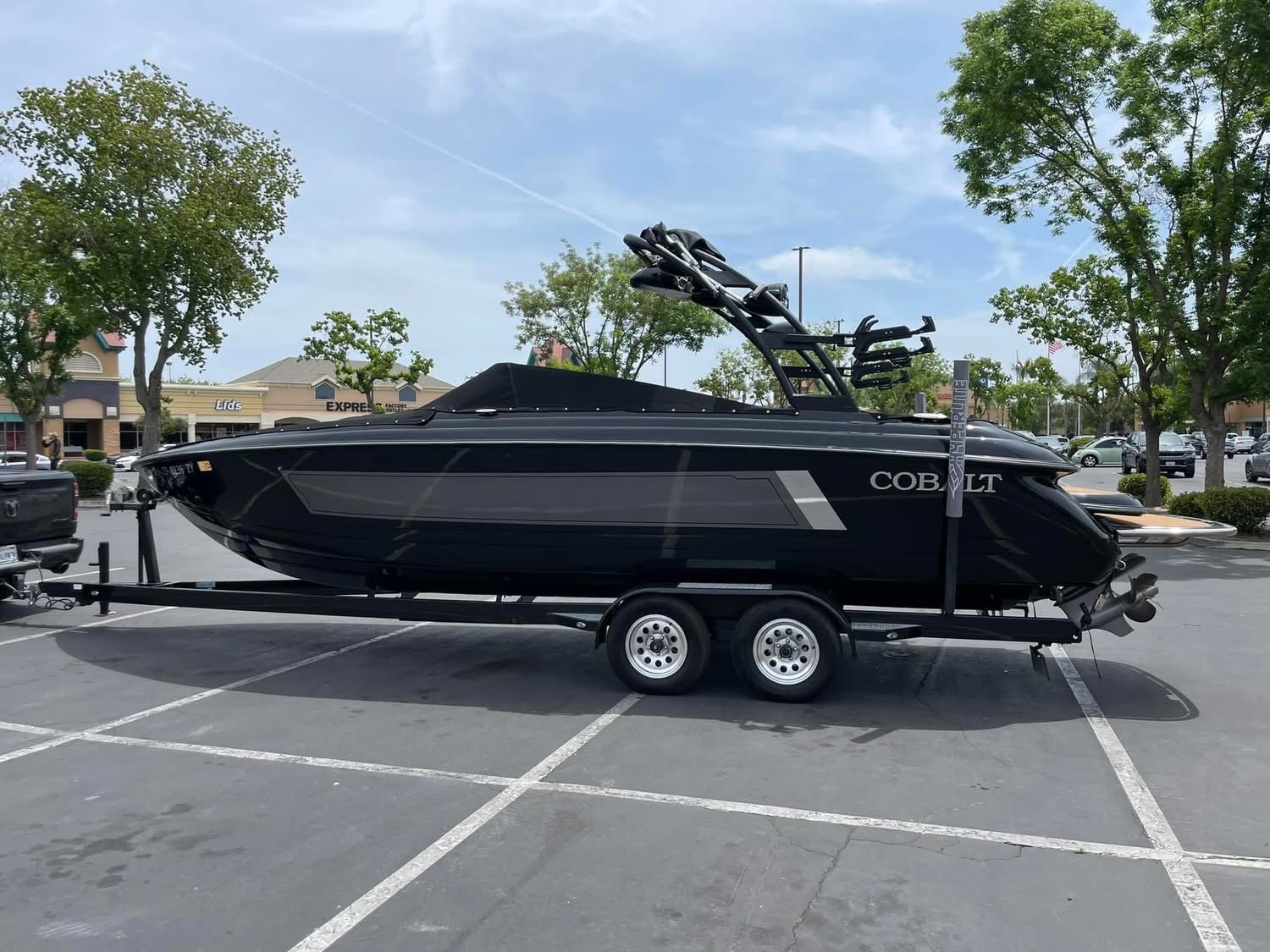 A black Cobalt speedboat on a double-axle trailer parked in a parking lot with trees and stores in the background.