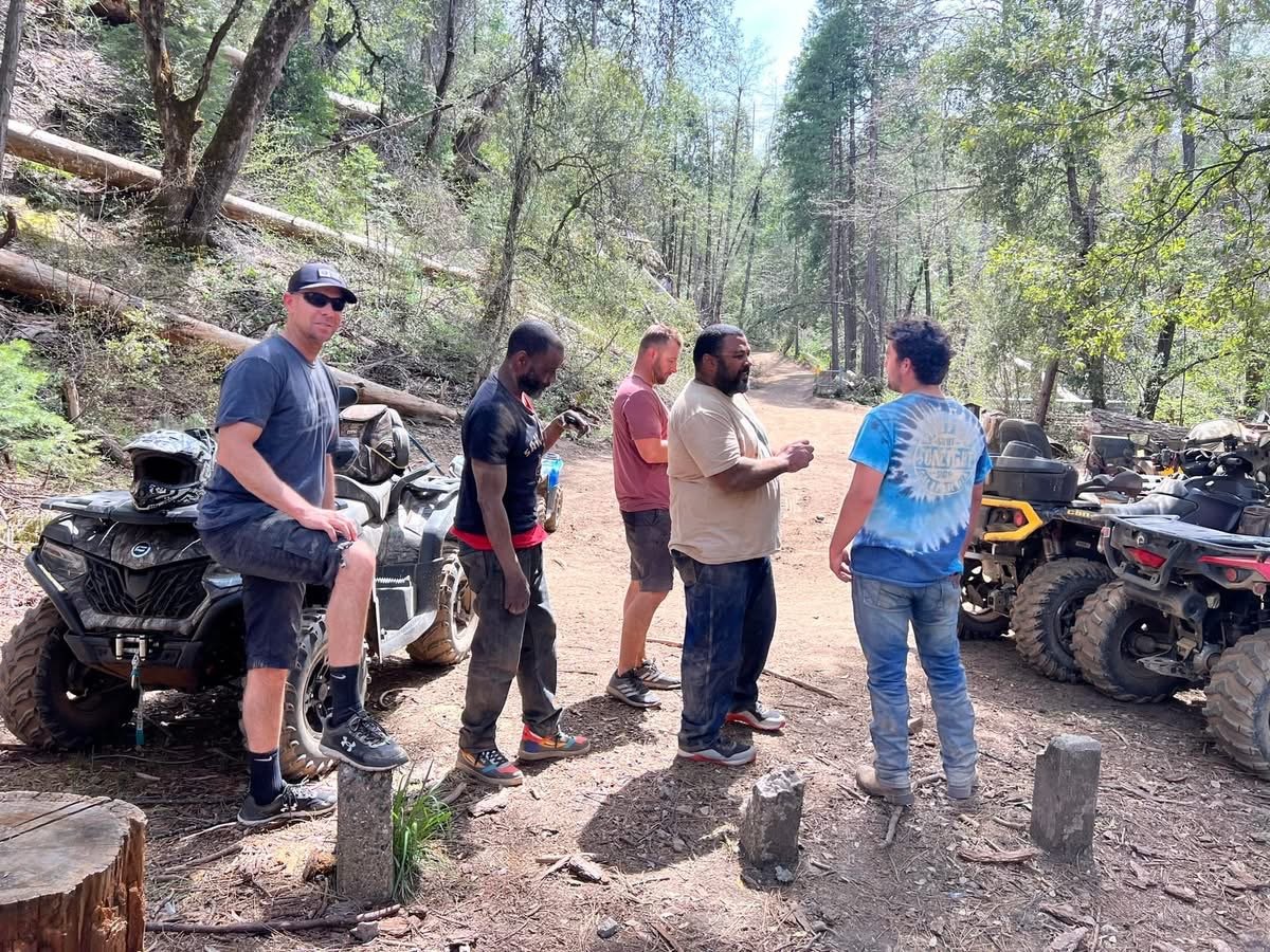 Group of five men chatting in a forested area with ATVs parked nearby.