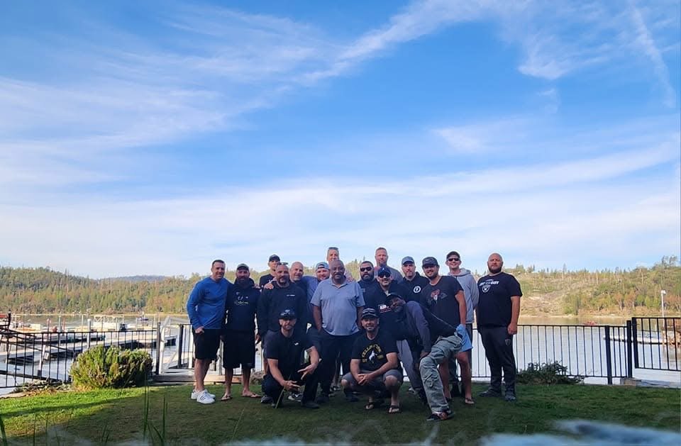 A group of men standing together outdoors near a body of water with trees and a blue sky in the background.