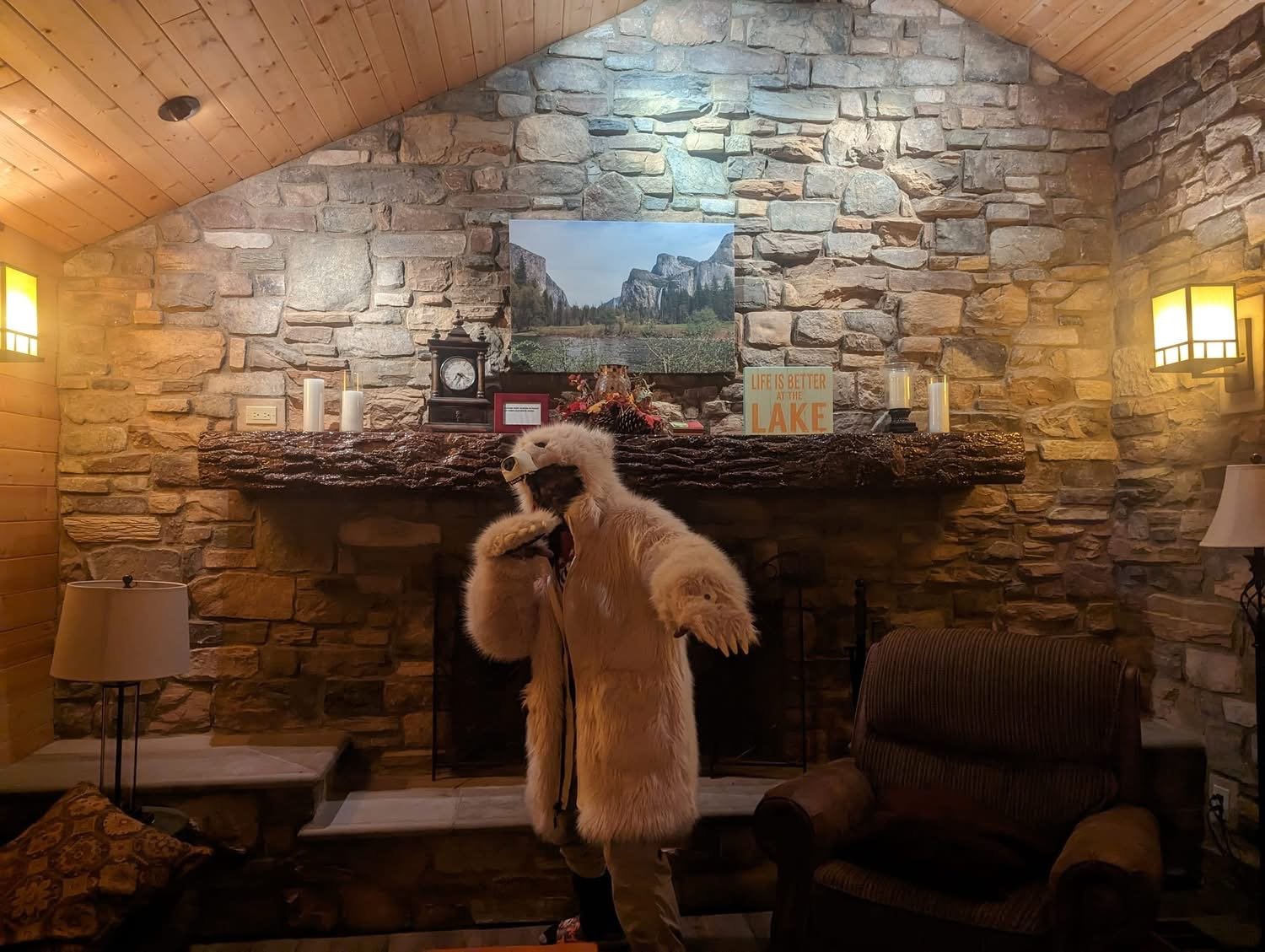 A person dressed in a polar bear costume making a yawning pose in front of a stone fireplace inside a cozy cabin living room with wooden walls and ceiling, with a large mountain landscape picture above the mantle, and lit lamps on either side.