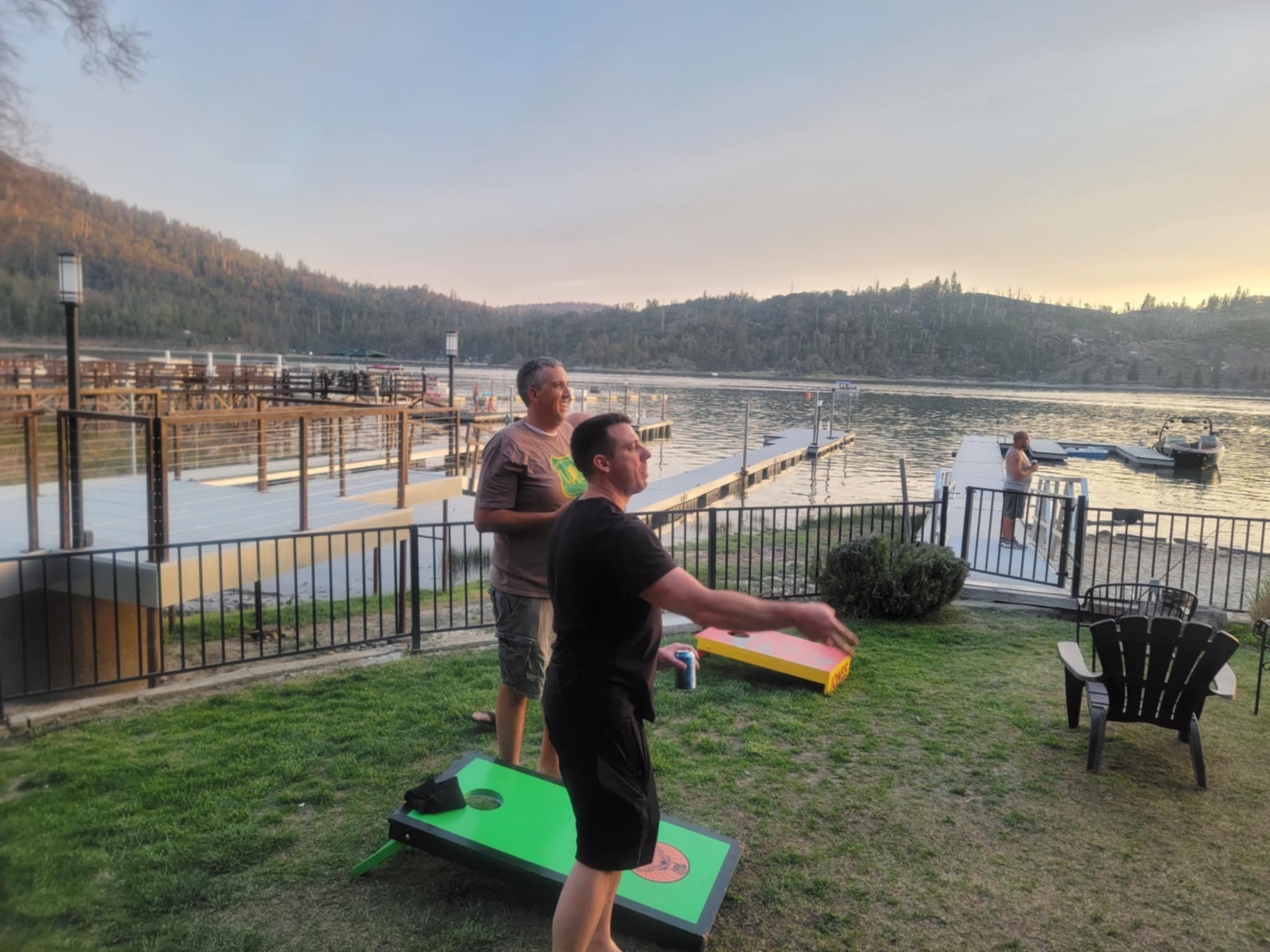 Two men playing cornhole near a lake during sunset, with a dock and boats in the background.