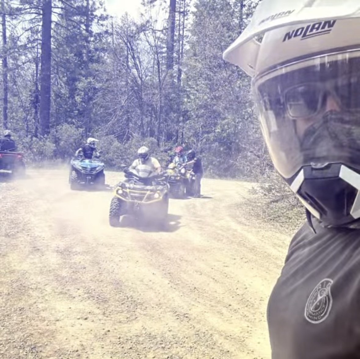 Person taking a selfie with a group of people riding all-terrain vehicles on a dirt trail in a wooded area, some wearing helmets.
