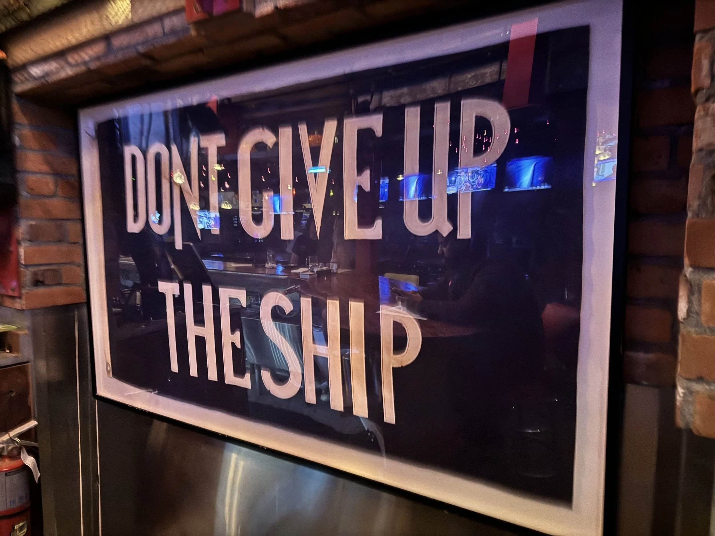 A sign with the words "DON'T GIVE UP THE SHIP" in large pink letters, hanging on a brick wall in a restaurant or bar. SFD Still F'cking Dangerous