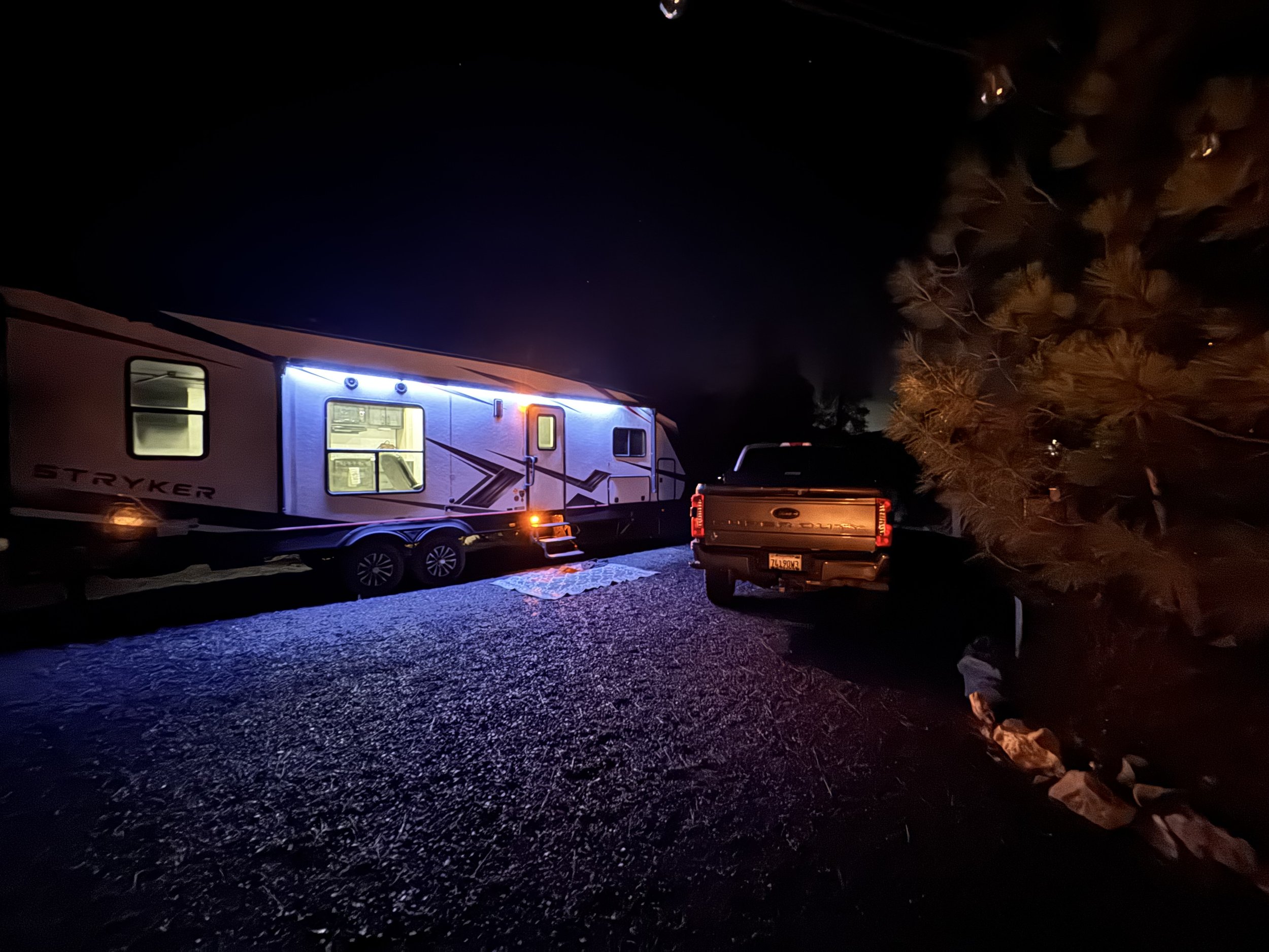 Nighttime scene of a parked RV with LED lights on, and a pickup truck beside it, surrounded by rocks and trees.