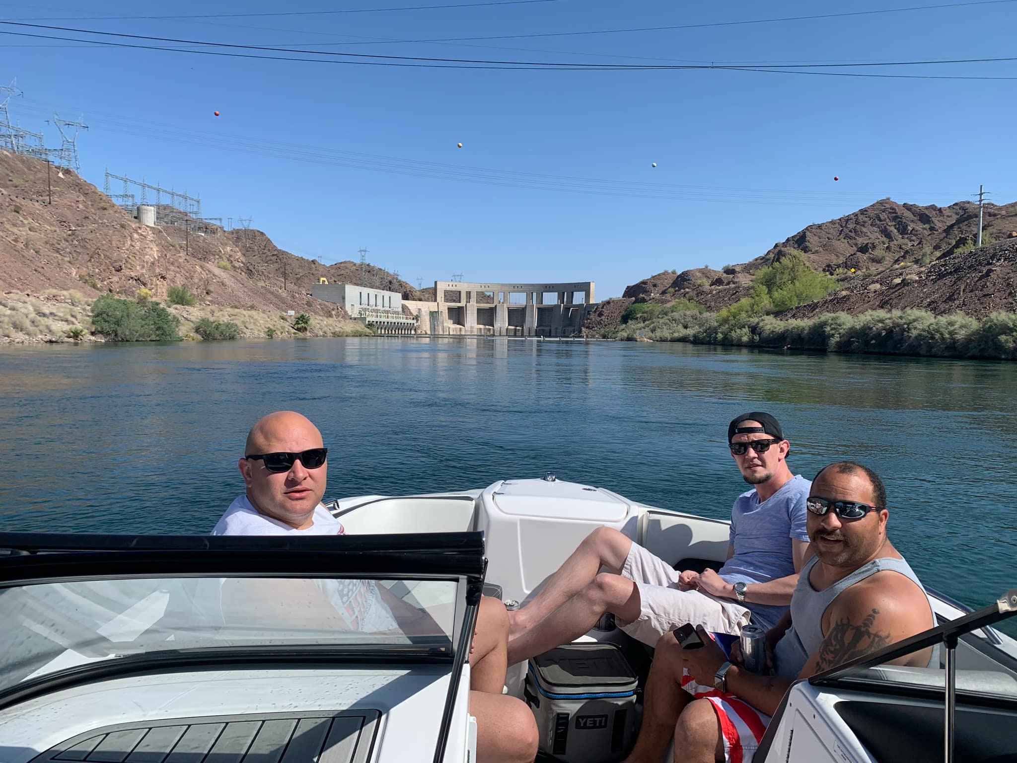 Three men sitting on a boat docked on a lake with mountains and a dam in the background, all wearing sunglasses and summer clothing, enjoying a sunny day.