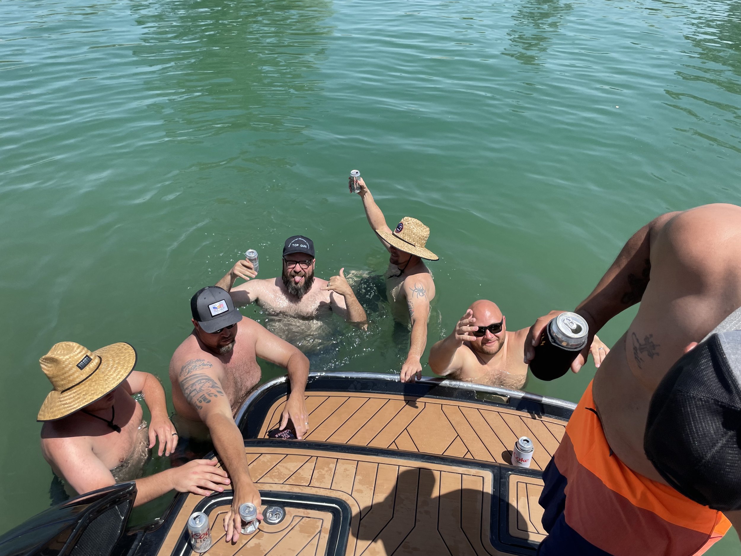 Group of five men, some with hats and tattoos, enjoying drinks in the water near a boat on a sunny day.
