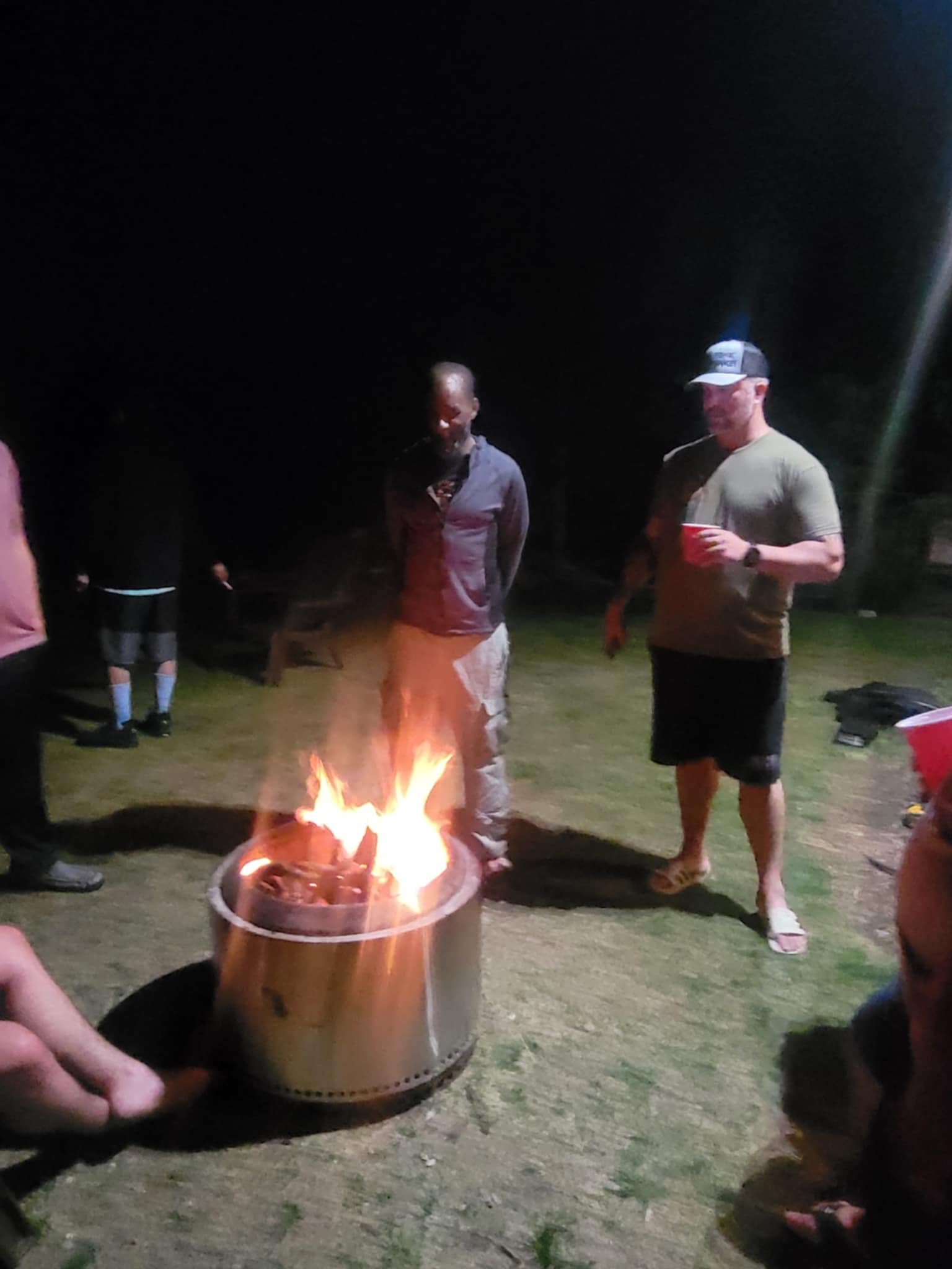 People gathered outdoors at night around a small fire in a metal fire pit, with two men in the center observing the flames. One man is holding a red cup, standing near another person, while others are partially visible in the background.