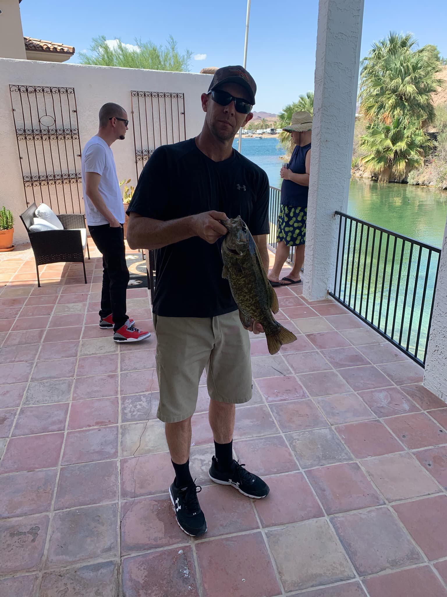 Man wearing black t-shirt, tan shorts, sunglasses, and black cap holding a large fish by the water on a patio. Two other people are standing in the background, one with a hat, and a man in a white shirt.